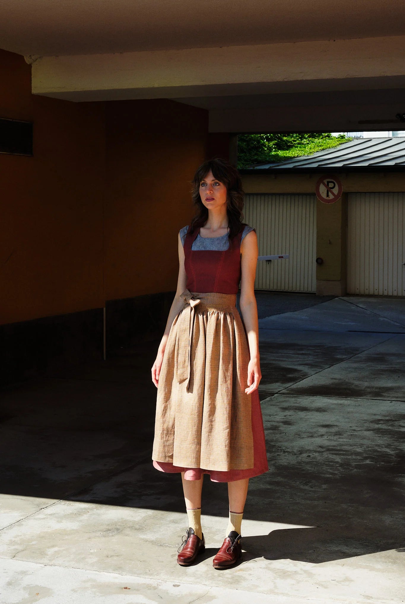 Model trägt Dirndl Ava in pinker Beere mit ocker-gestreifter Schürze – steht regungslos in einer Garageneinfahrt, halb im Schatten, halb in der Sonne, urban-minimalistisch inszeniert von Zirbenfux