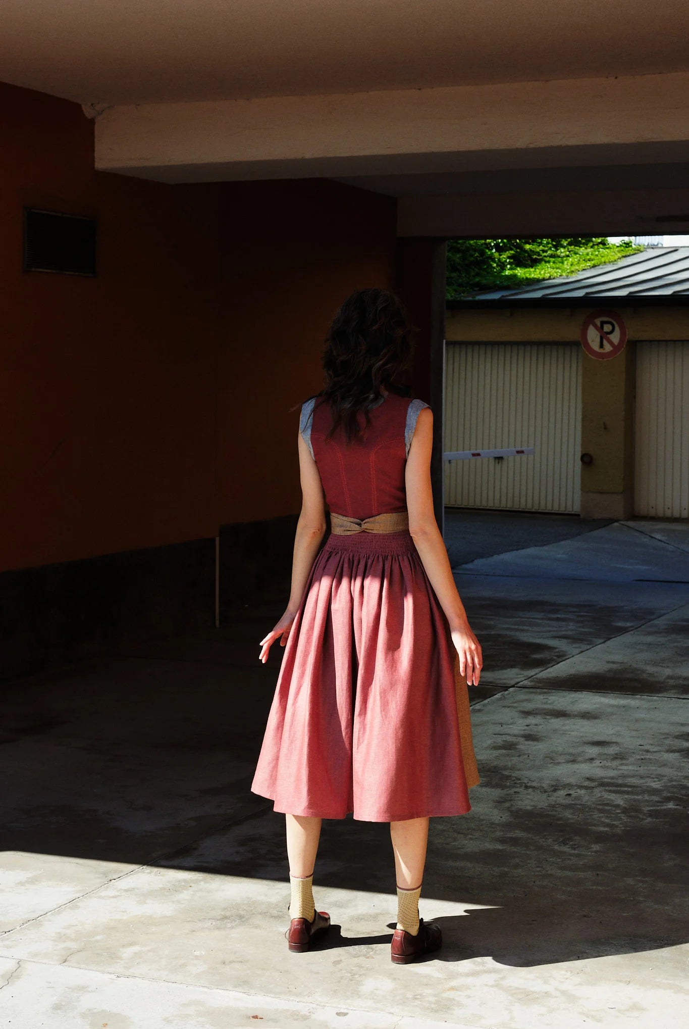 Model trägt Dirndl Ava in pinker Beere mit ocker-gestreifter Schürze – steht mit dem Rücken zur Kamera in einer Garageneinfahrt, halb im Schatten, halb im Sonnenlicht, ruhig und urban inszeniert von Zirbenfux
