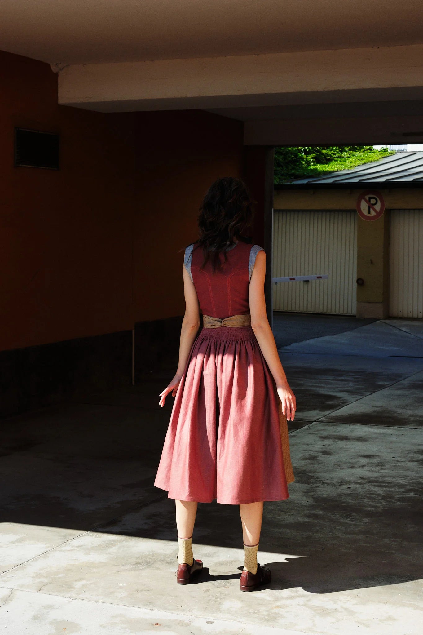 Model trägt Dirndl Ava in pinker Beere mit ocker-gestreifter Schürze – steht mit dem Rücken zur Kamera in einer Garageneinfahrt, halb im Schatten, halb im Sonnenlicht, ruhig und urban inszeniert von Zirbenfux