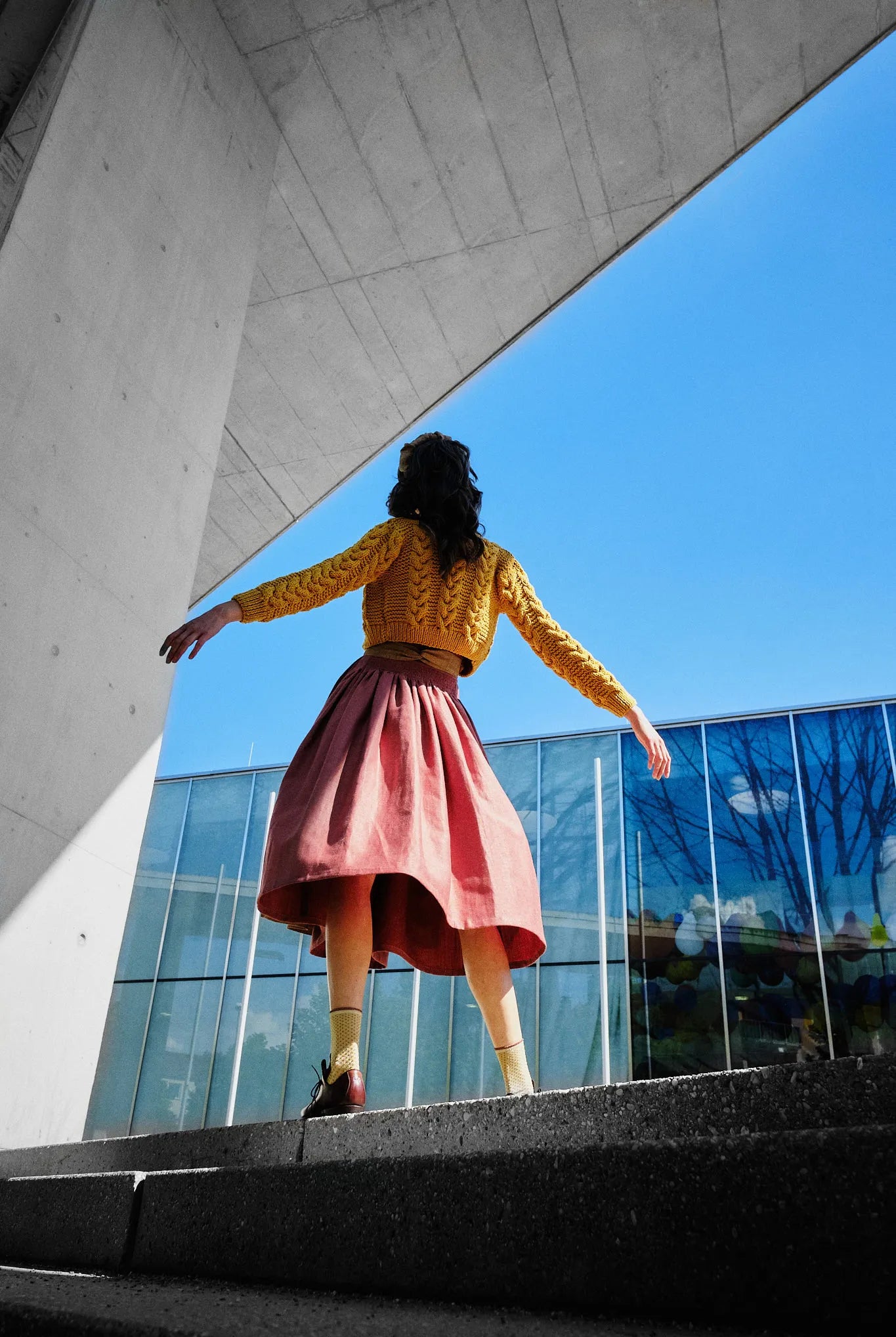 Dirndl Ava in Pinke Beere aus Leinen mit ocker gestreifter Schürze und Strickjacke, Rückansicht mit Perspektive vor moderner Fassade an Treppenabsatz mit seitlich ausgestreckten Armen und weit schwingendem Rock, kraftvoll inszeniert von Zirbenfux.