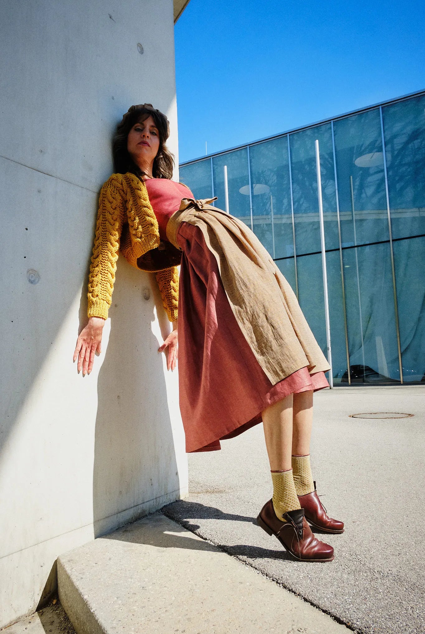 Dirndl Ava in Pinke Beere aus Leinen mit ocker gestreifter Schürze und Strickjacke, Model überstreckt nach hinten an eine Säule gelehnt vor modernem urbanem Gebäude, ausdrucksstark inszeniert von Zirbenfux.