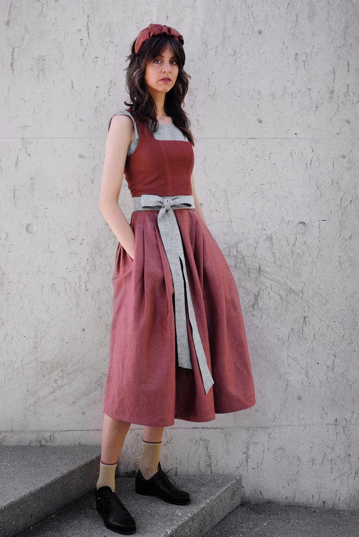 Dirndl Ava in Pinke Beere aus Leinen mit blaugrauem Schürzenband, Model klar und gerade vor einer modernen Waschbetonwand stehend, Hände locker in den Rocktaschen, gelassener Blick in die Kamera, ruhig inszeniert von Zirbenfux.