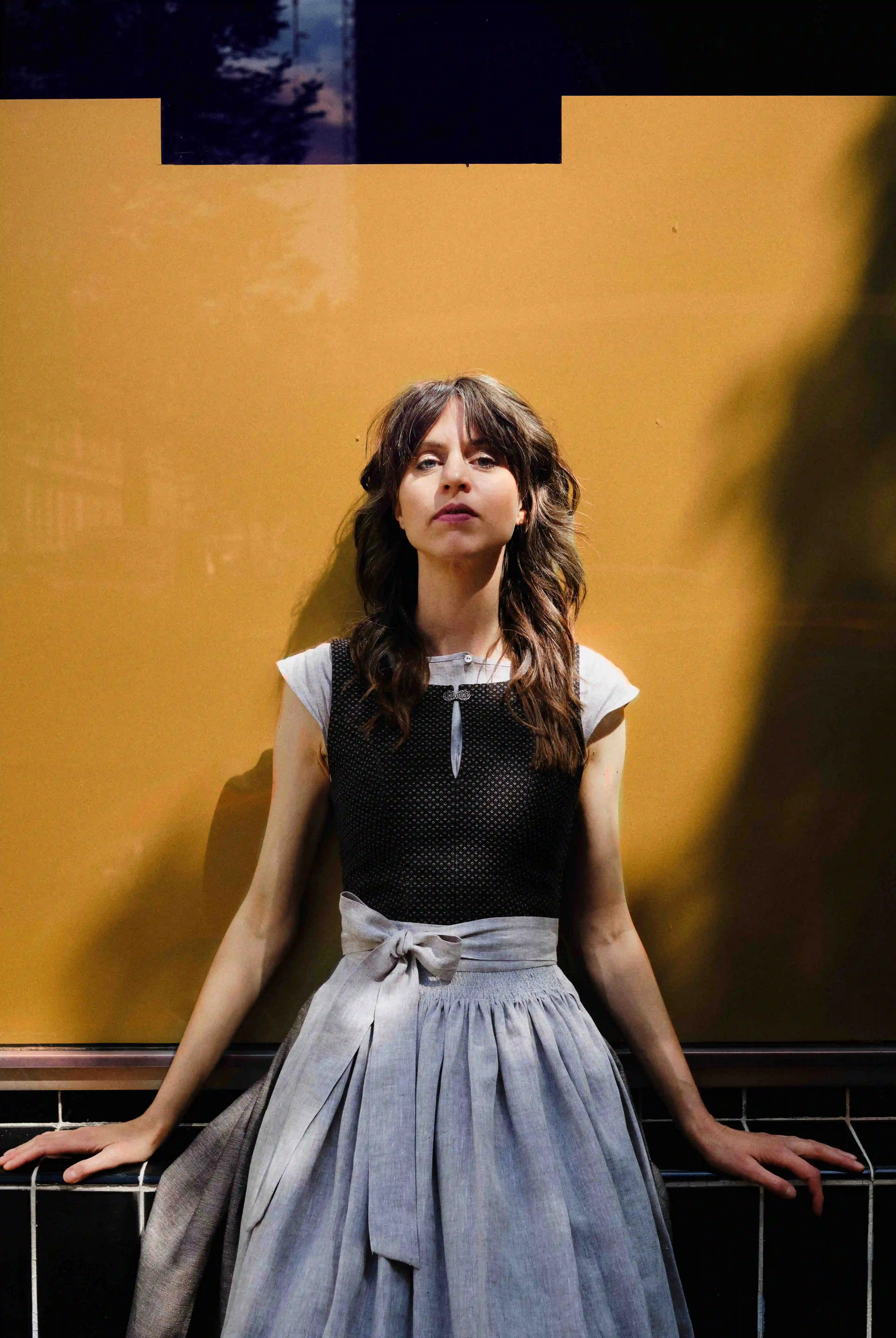 Model trägt Dirndl Marlene in Schwarz-Natur mit Leinenschürze – sitzt vor goldenem Fenster, Hände locker zu beiden Seiten abgelegt, direkter, kühler Blick in die Kamera, Licht und Schattenspiel, stilvoll inszeniert von Zirbenfux