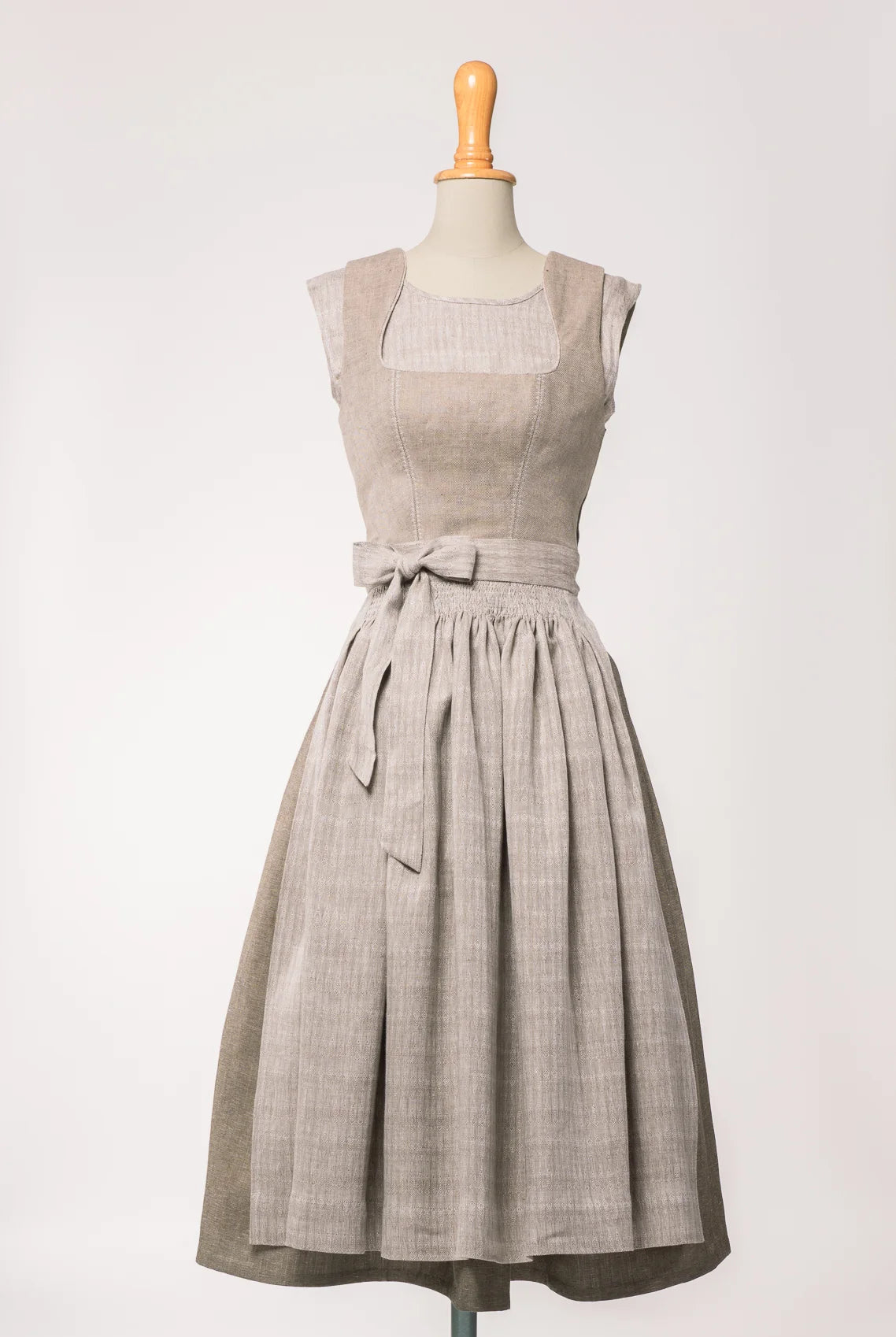 Dirndlschürze aus naturfarbenem Bio-Leinen mit eingewebtem Muster, kombiniert mit Mieder Ava in hellem Naturton und Rock Marlene in goldbraun – Frontansicht auf Büste, handgefertigt von Zirbenfux