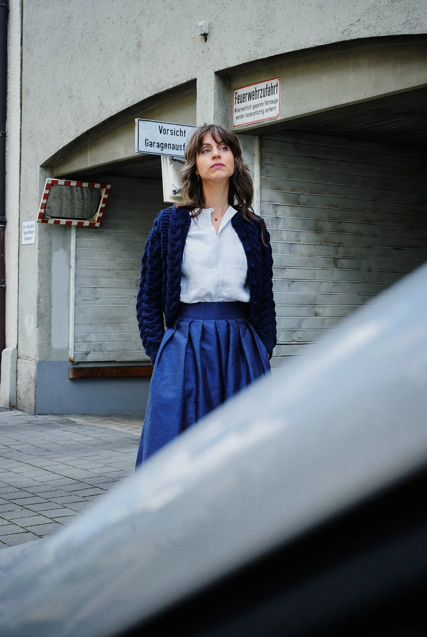 Hosenrock Audrey mit Bluse Romy und Strickjacke im Set in Blau aus Leinen, Model stehend in einer urbanen Garageneinfahrt mit Blick in die Ferne, Auto im Anschnitt im Vordergrund, ruhig interpretiert von Zirbenfux.