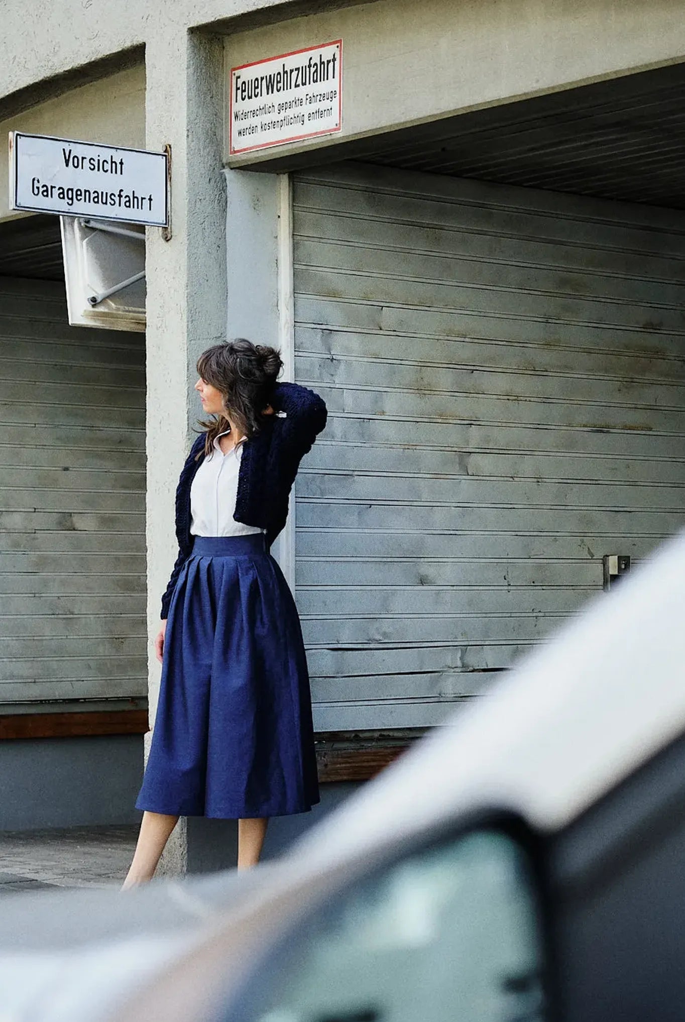 Hosenrock Audrey mit Bluse Romy und Strickjacke im Set in Blau aus Leinen, Model stehend an einer urbanen Garageneinfahrt mit abgewandtem Blick, Auto im Anschnitt im Vordergrund, klar inszeniert von Zirbenfux.