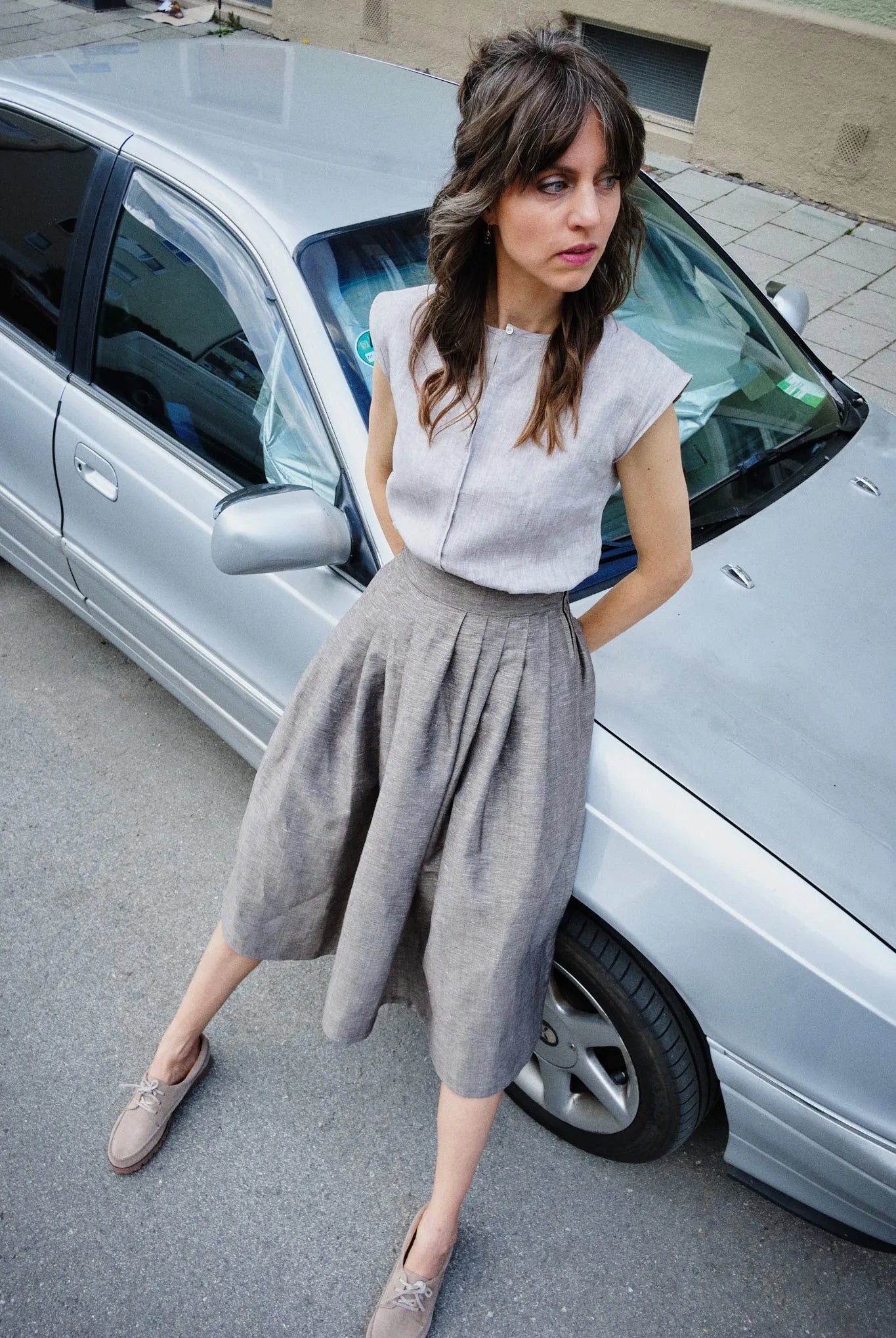 Model trägt Trachtenrock Marlene aus goldbrauner Leinen-Schurwolle-Mischung und Bluse Tilda aus naturfarbenem Bio-Leinen – lehnt lässig an silbernem Auto in urbanem Umfeld, Blick seitlich aus dem Bild, zeitgemäß interpretiert von Zirbenfux