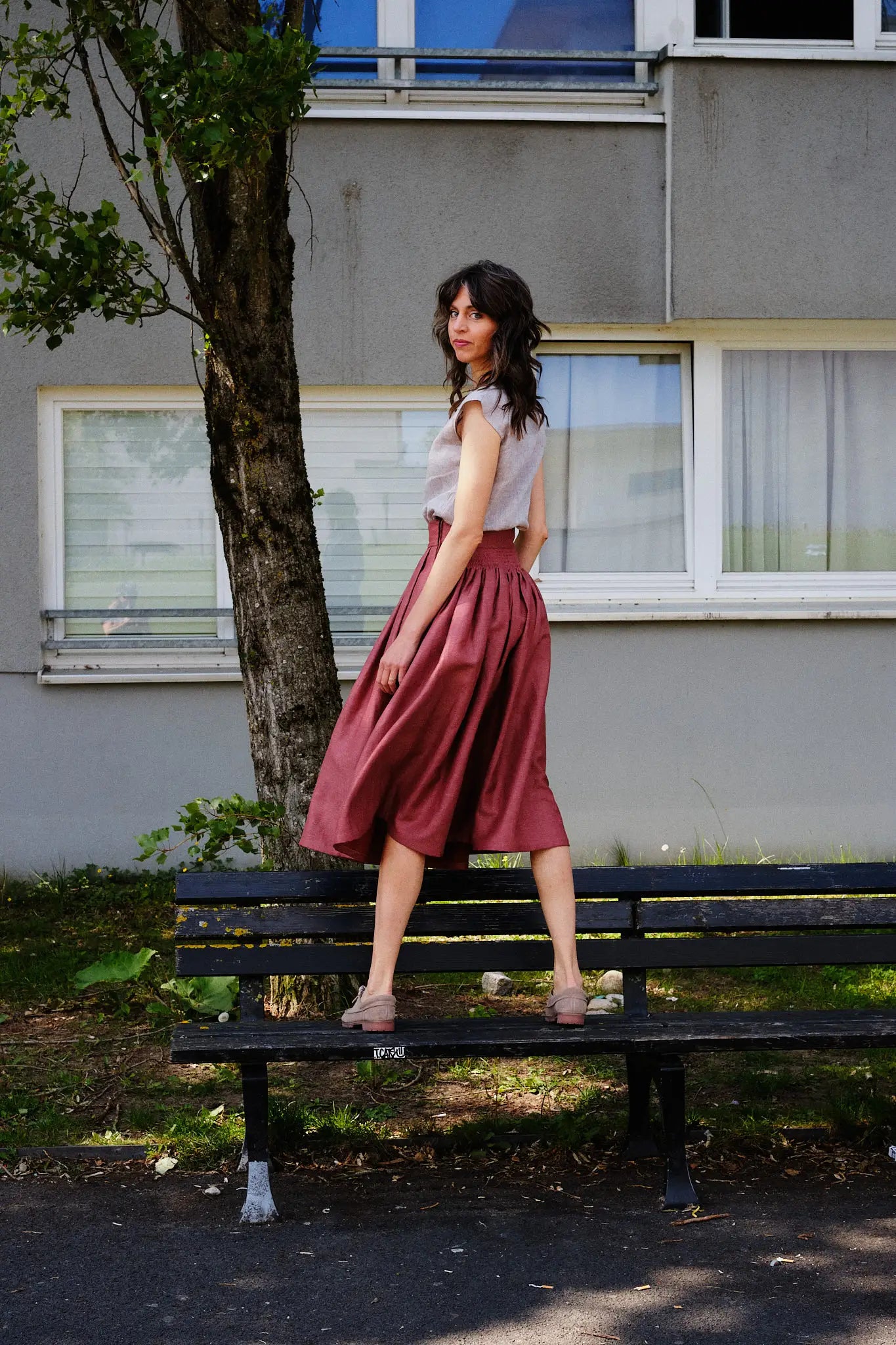 Trachtenrock Marlene in Pinke Beere mit Bluse TILDA im Set aus Leinen in Natur, Model gerade auf einer Bank stehend mit dem Rücken zum Betrachter und über die Schulter in die Kamera blickend, ruhig inszeniert von Zirbenfux.