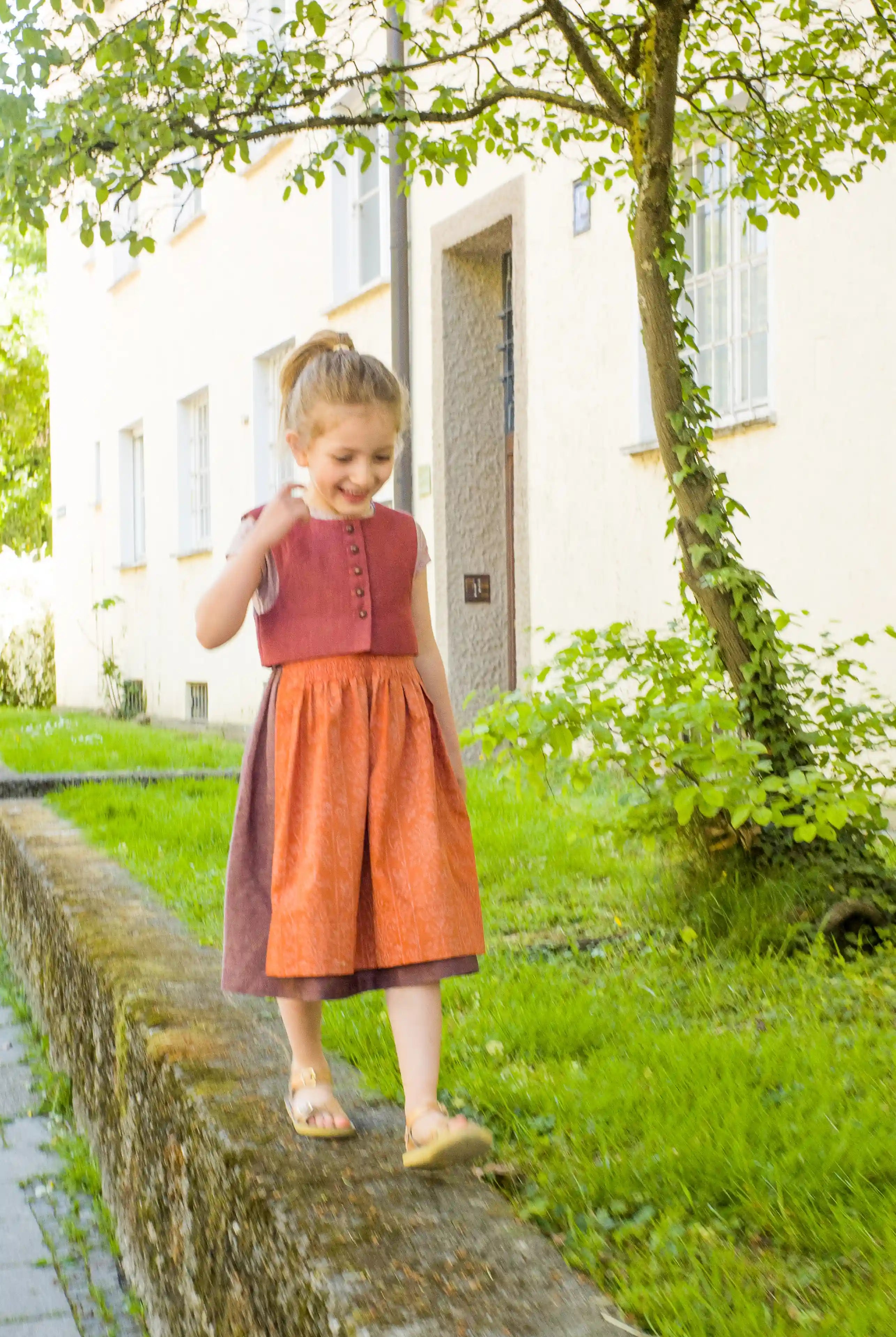 Kinderdirndl in pinker Beere mit terrakottafarbener Schürze – balancierend auf einem Mäuerchen vor urbaner Häuserfront, modern inszeniert von Zirbenfüxlein