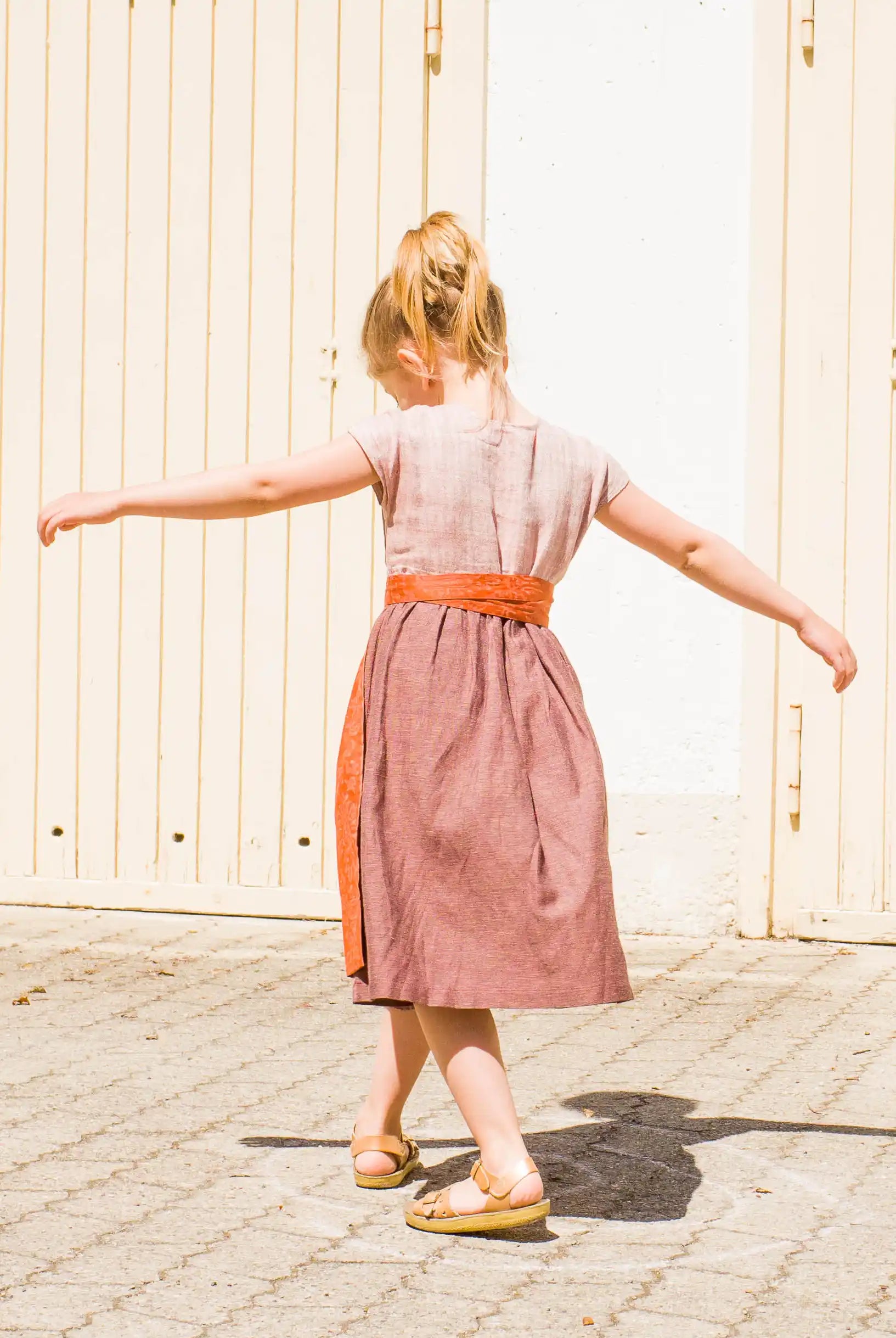 Kinderdirndl in Rosenholz mit terrakottafarbener Schürze – Rückansicht beim Drehen vor Garagentor, Arme weit zur Seite gestreckt, strahlende Sonne auf Asphalt, lebendig und frei inszeniert von Zirbenfüxlein