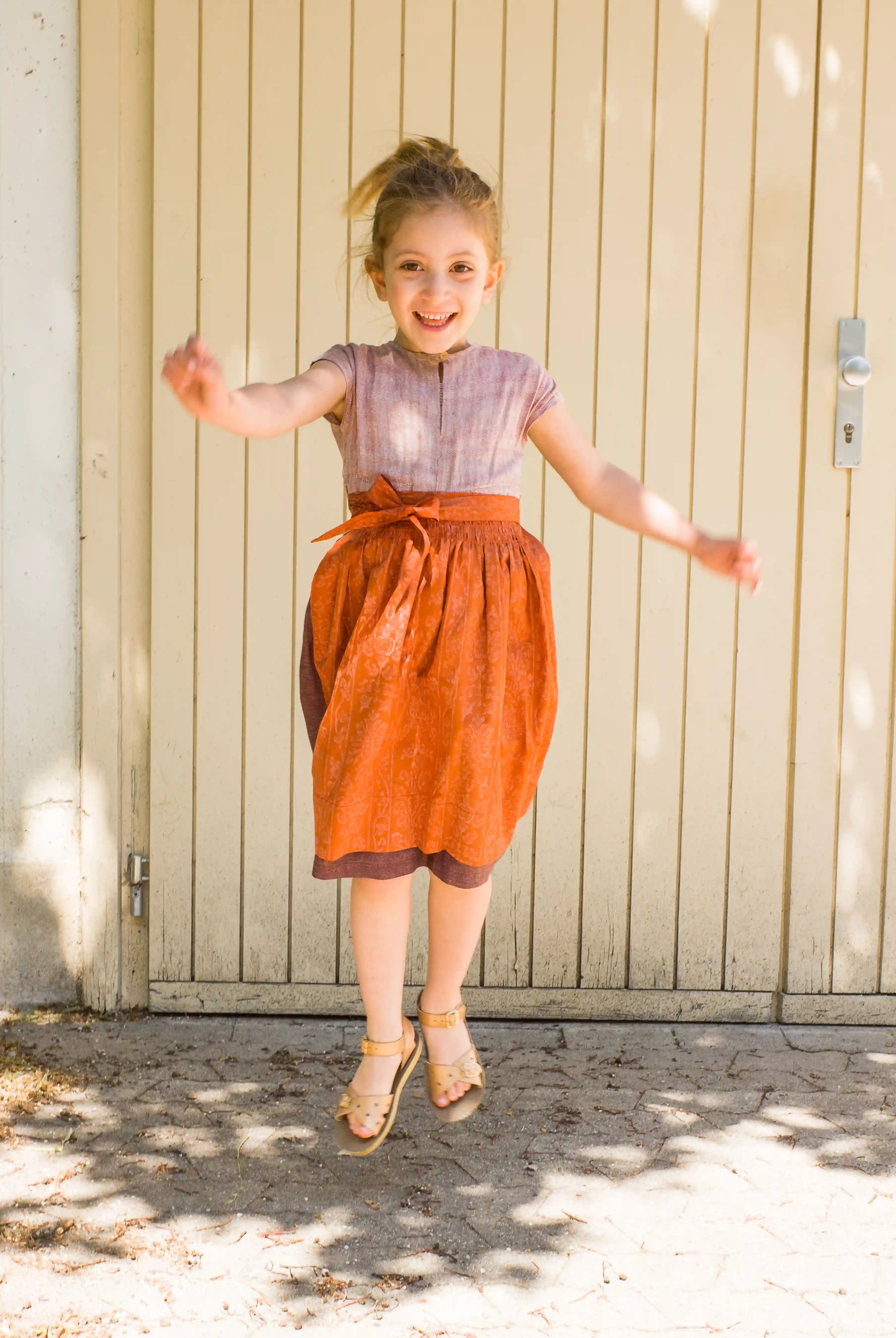Kinderdirndl in Rosenholz mit terrakottafarbener Schürze – springend und lachend vor einem Garagentor, lebendige Szene mit Schattenspiel, spielerisch inszeniert von Zirbenfüxlein