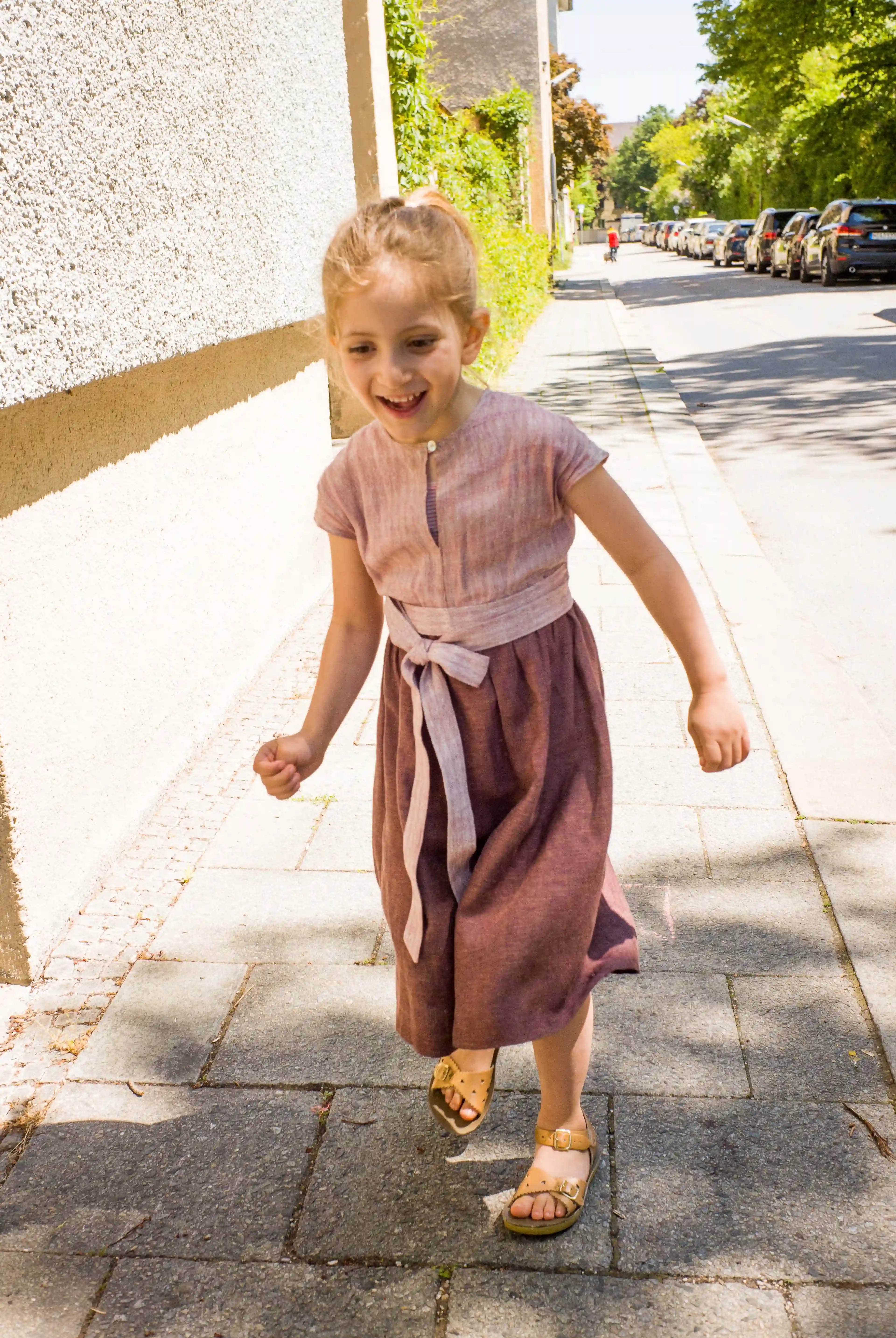 Kinderdirndl in Rosenholz mit Schürzenband – Kind läuft auf dem Gehweg entlang einer Straße im urbanen Setting, fröhlich und modern inszeniert von Zirbenfüxlein
