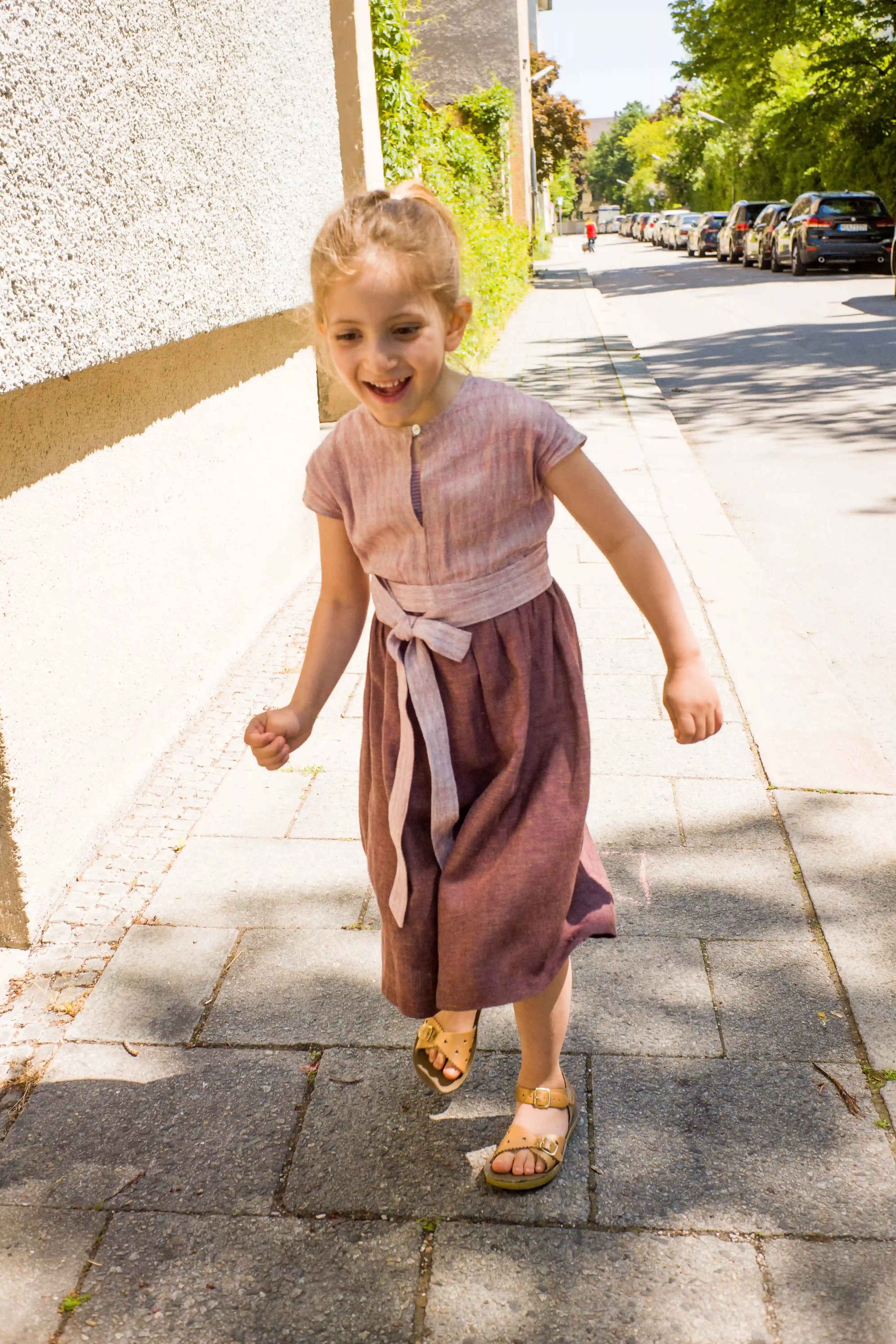Kinderdirndl in Rosenholz mit Schürzenband – Kind läuft auf dem Gehweg entlang einer Straße im urbanen Setting, fröhlich und modern inszeniert von Zirbenfüxlein