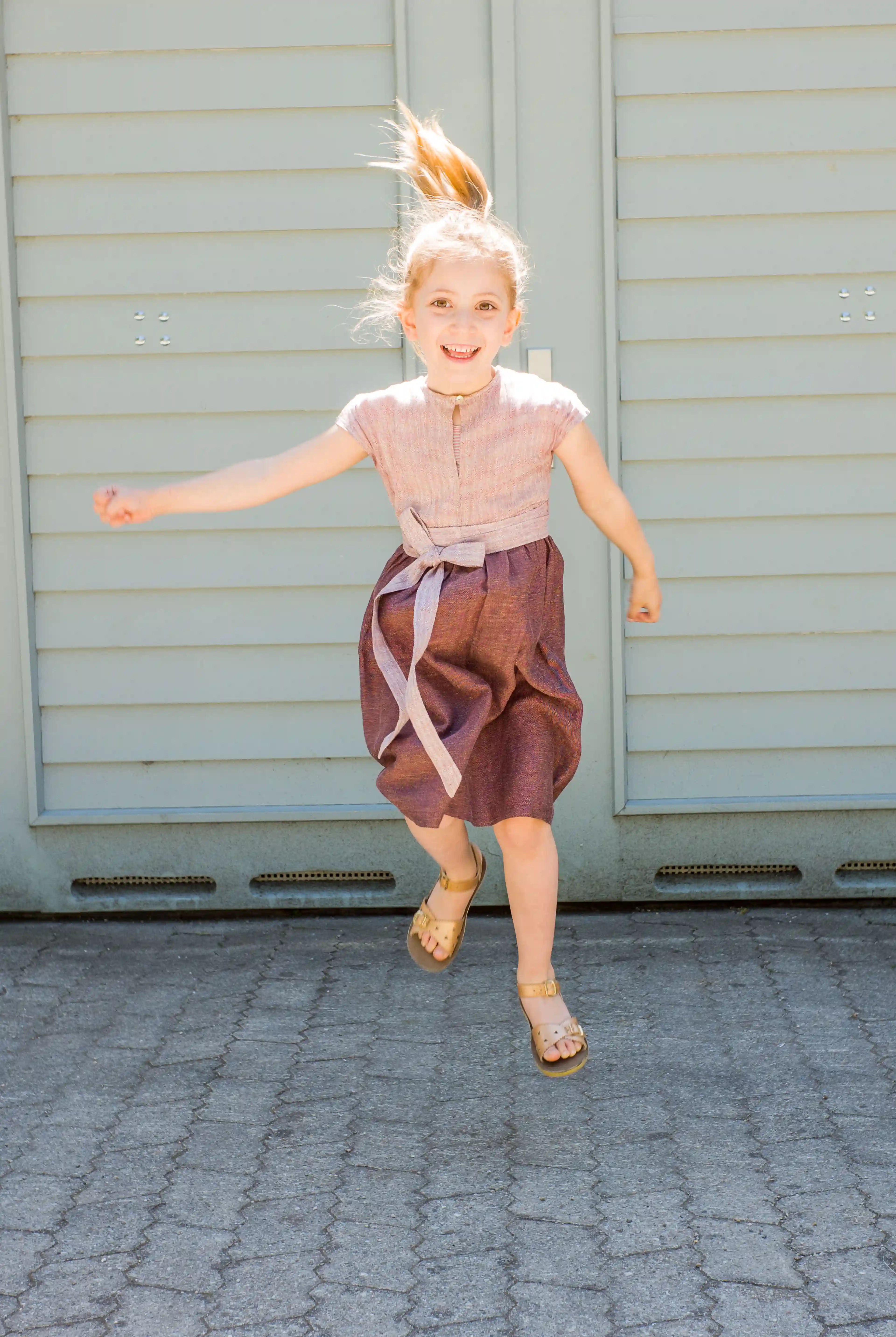 Kinderdirndl in Rosenholz mit Schürzenband – fröhlich nach oben springend vor einem Garagentor im Sonnenschein, dynamisch und natürlich inszeniert von Zirbenfüxlein