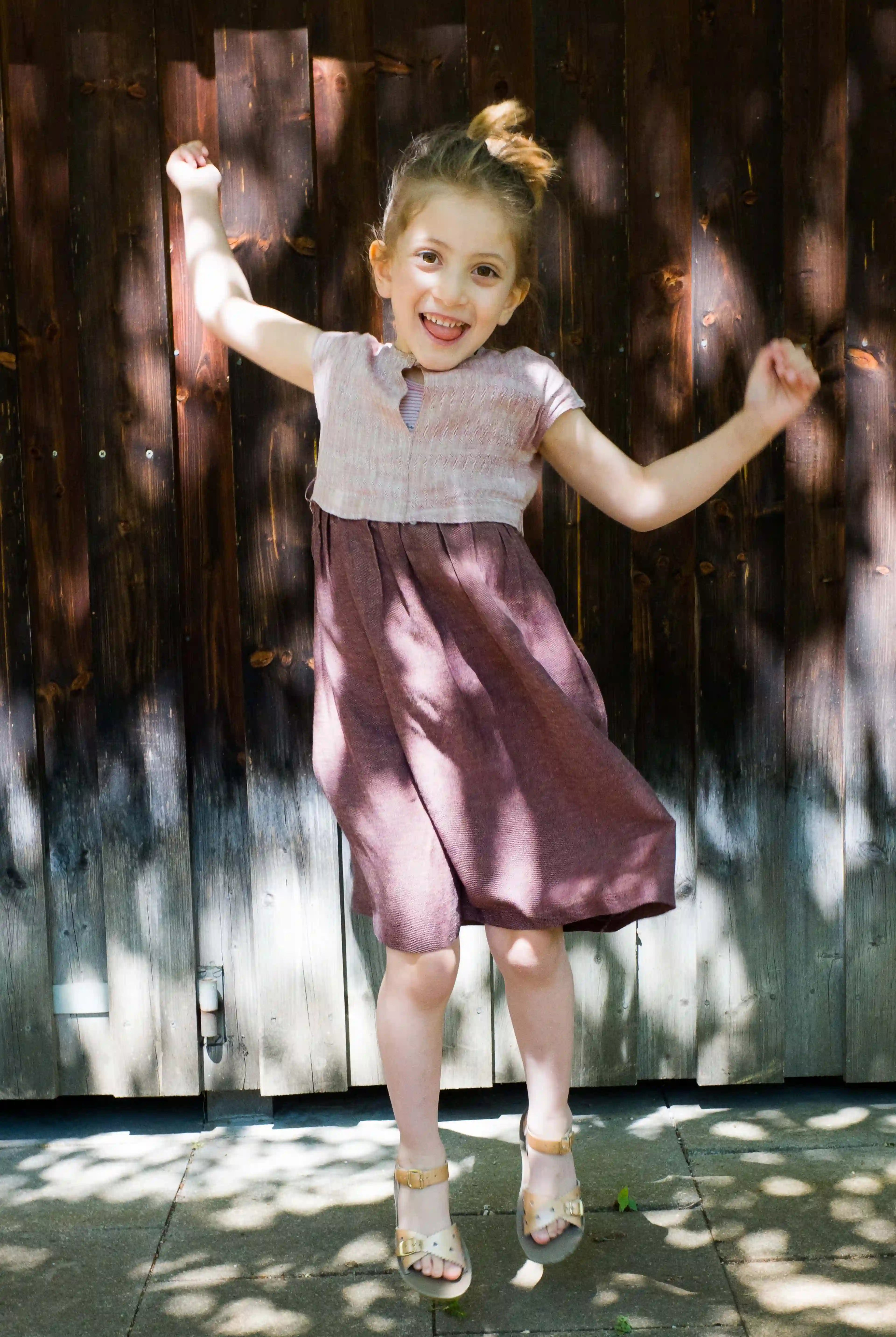 Kinderdirndl in Rosenholz – fröhlich springend vor Altholzwand auf Asphalt, mit kontrastreichem Schattenspiel, spielerisch inszeniert von Zirbenfüxlein