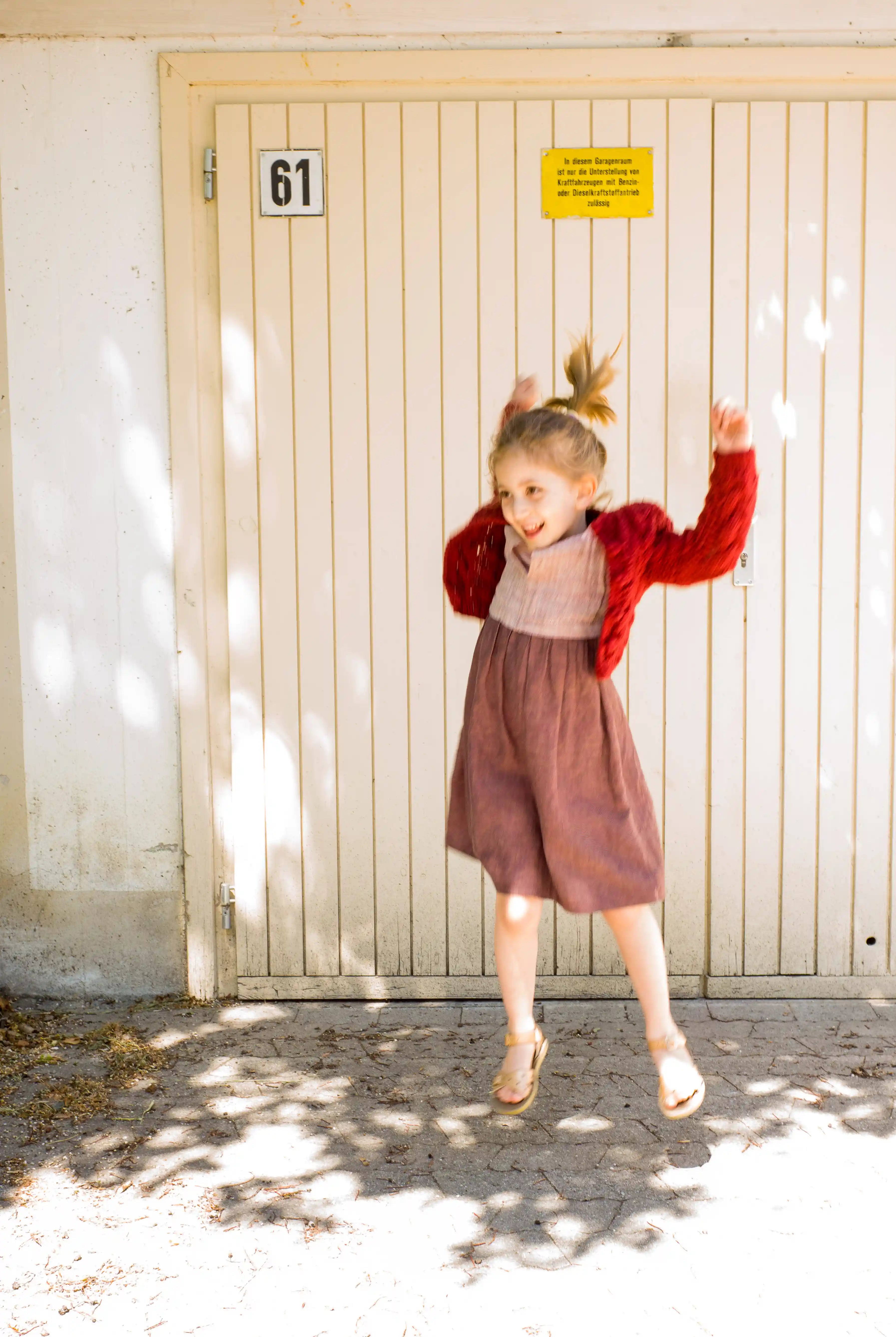 Kinderdirndl in Rosenholz mit kirschroter Strickjacke – fröhlich springend, dynamische Szene mit Unschärfe im Bild, spontan inszeniert von Zirbenfüxlein