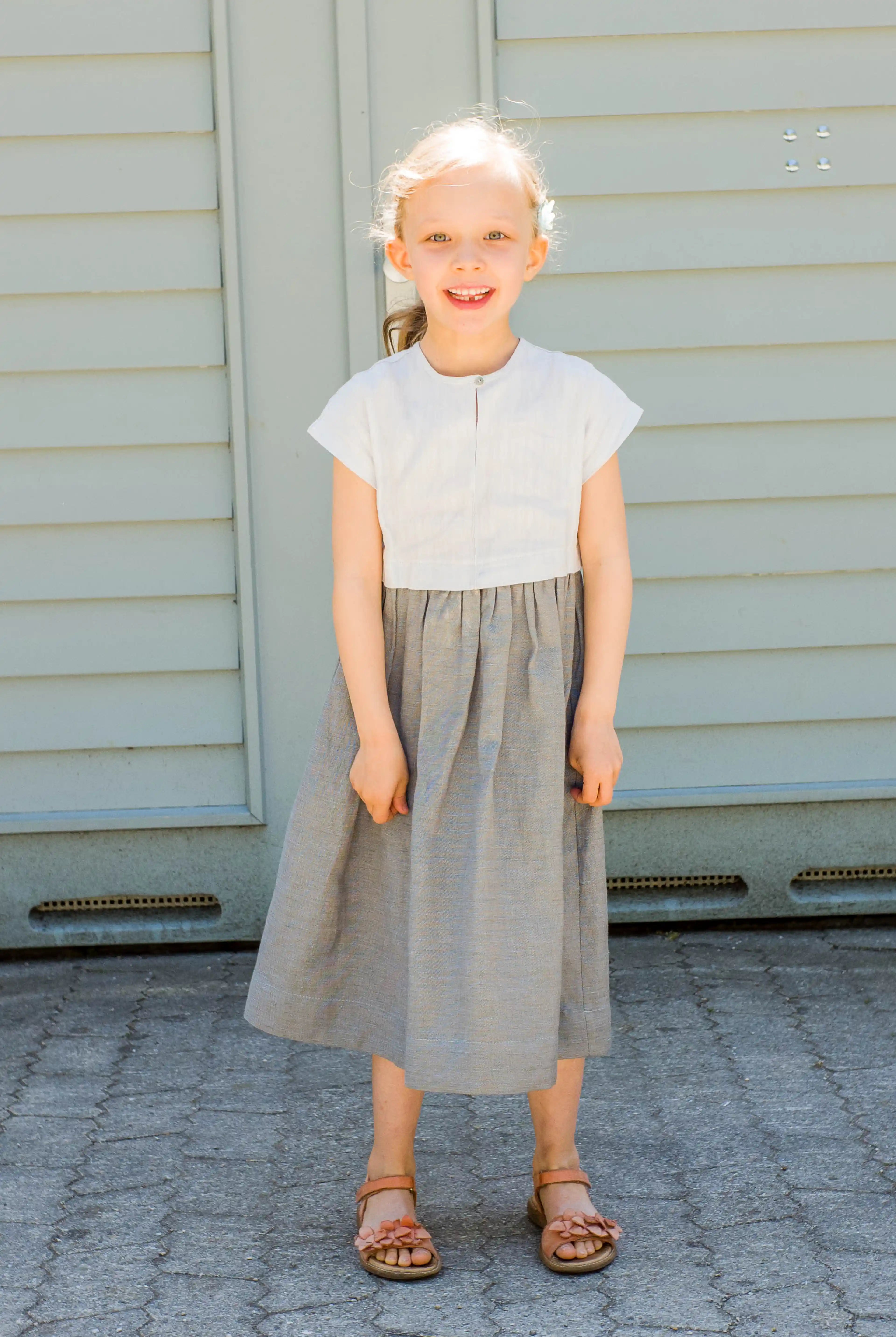 Kinderdirndl in Saphirblau – frontal vor einem Garagentor in heller Sonne stehend, fröhlich und modern inszeniert von Zirbenfüxlein
