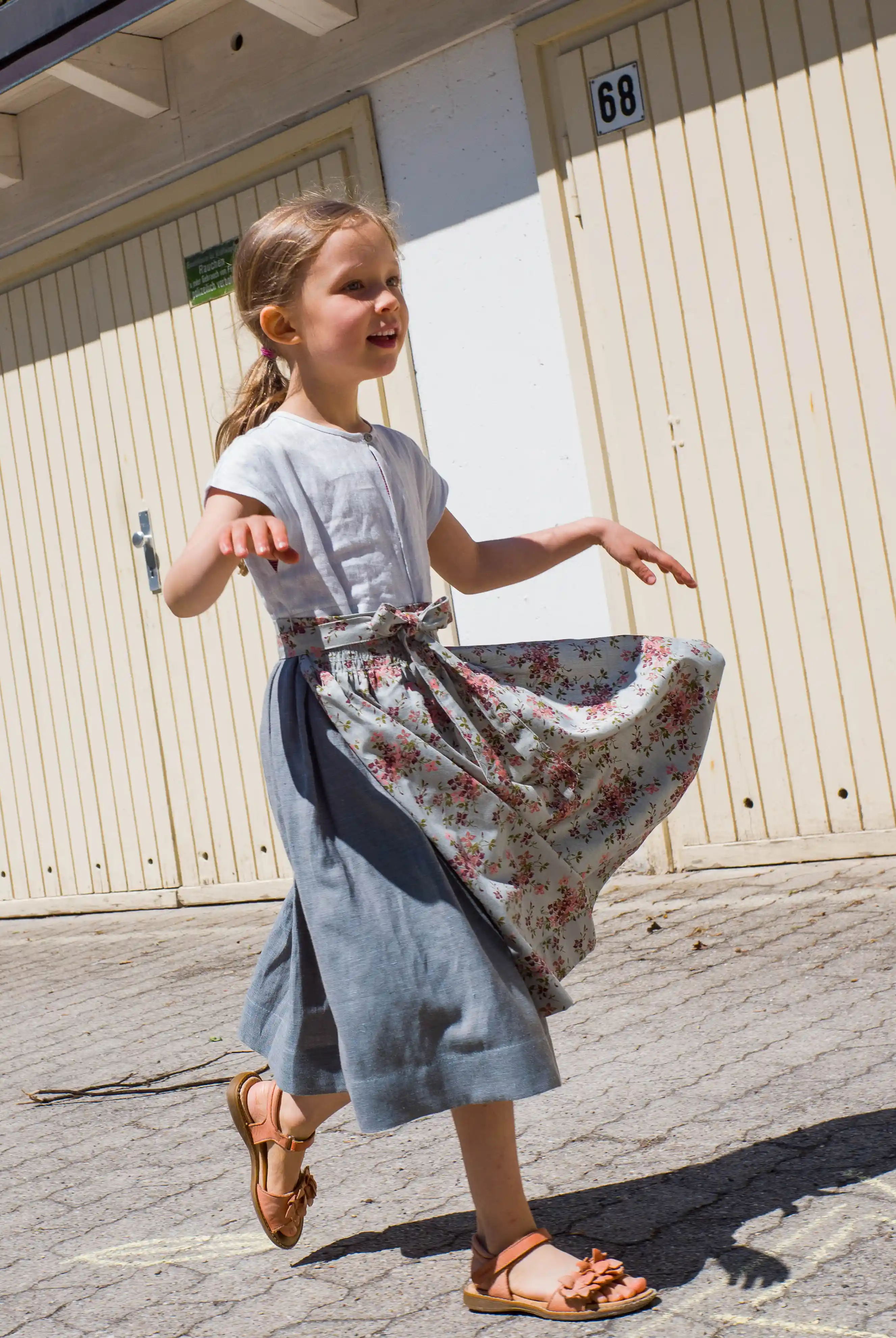 Zirbenfüxlein Kinderdirndl Saphir aus Leinen mit hellblauer Schürze mit Blumenmuster, Kind läuft entlang einer Garageneinfahrt, Schürze in Bewegung vor urbanen Garagentoren, lebendig umgesetzt von Zirbenfüxlein.