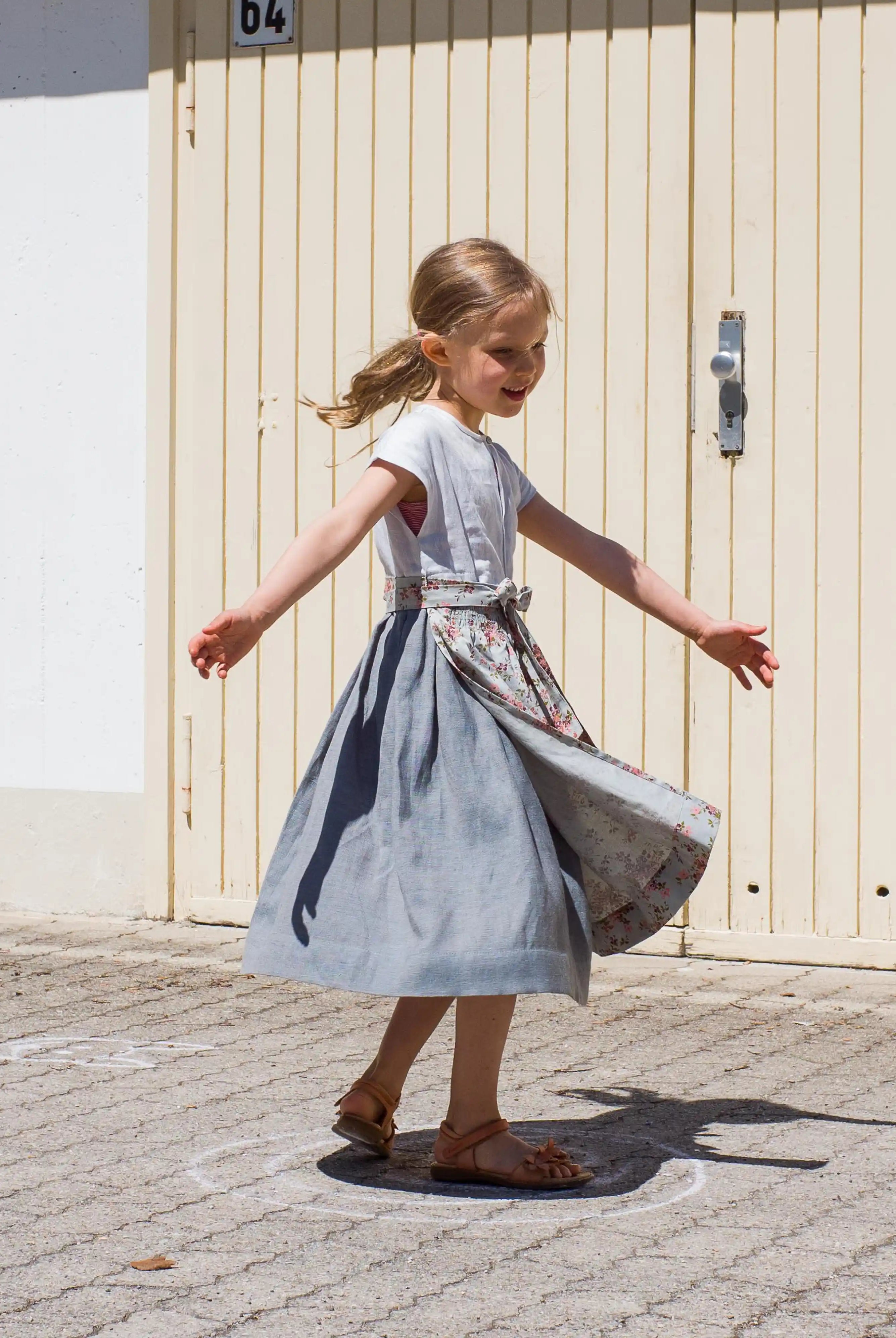 Zirbenfüxlein Kinderdirndl Saphir aus Leinen mit hellblauer Schürze mit Blumenmuster, schwungvolle Drehung mit ausgestreckten Armen in Frontansicht vor urbanen Garagentoren, lebendig umgesetzt von Zirbenfüxlein.