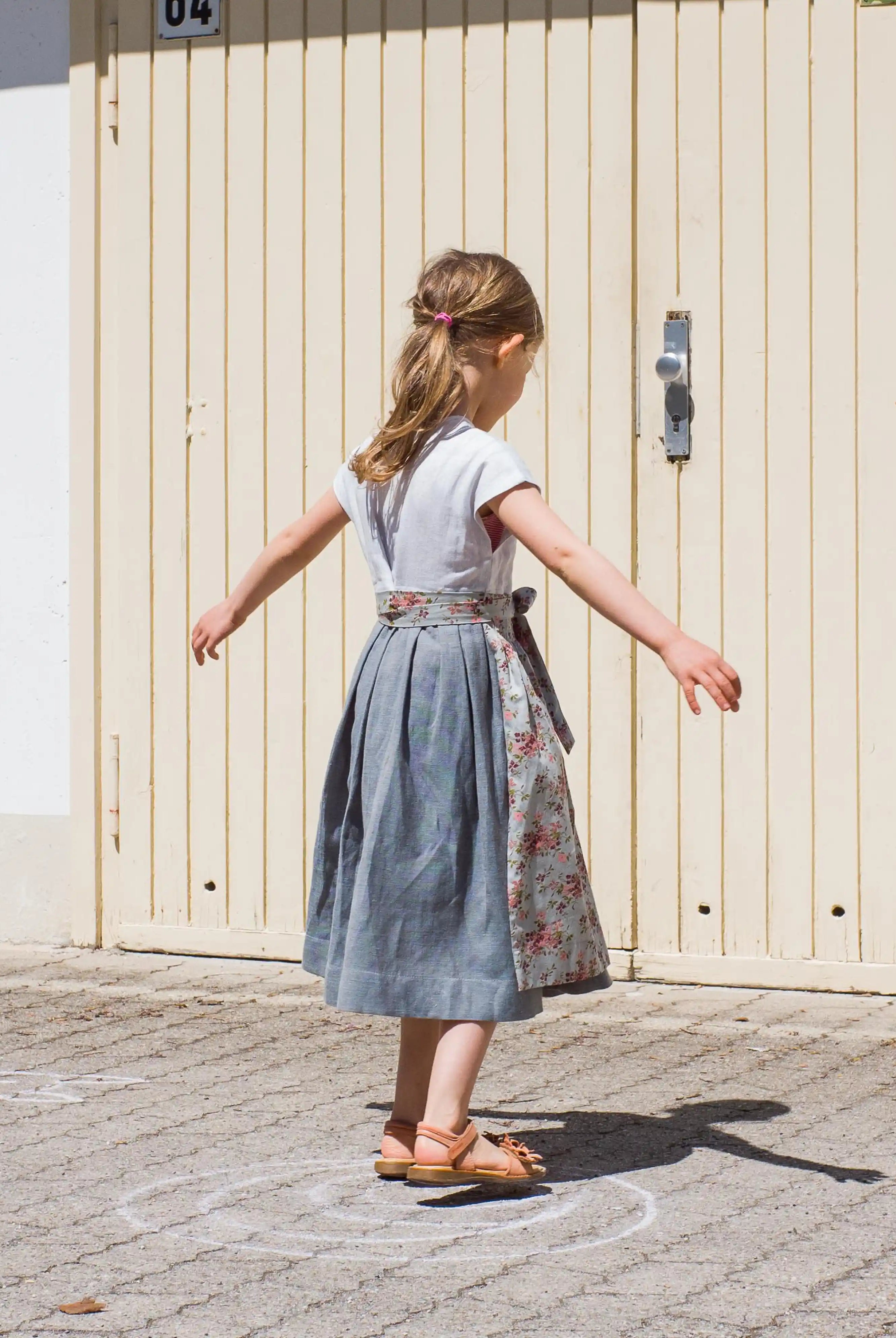 Zirbenfüxlein Kinderdirndl Saphir aus Leinen mit hellblauer Schürze mit Blumenmuster, Kind in Drehbewegung in Rückansicht vor urbanen Garagentoren, lebendig umgesetzt von Zirbenfüxlein.