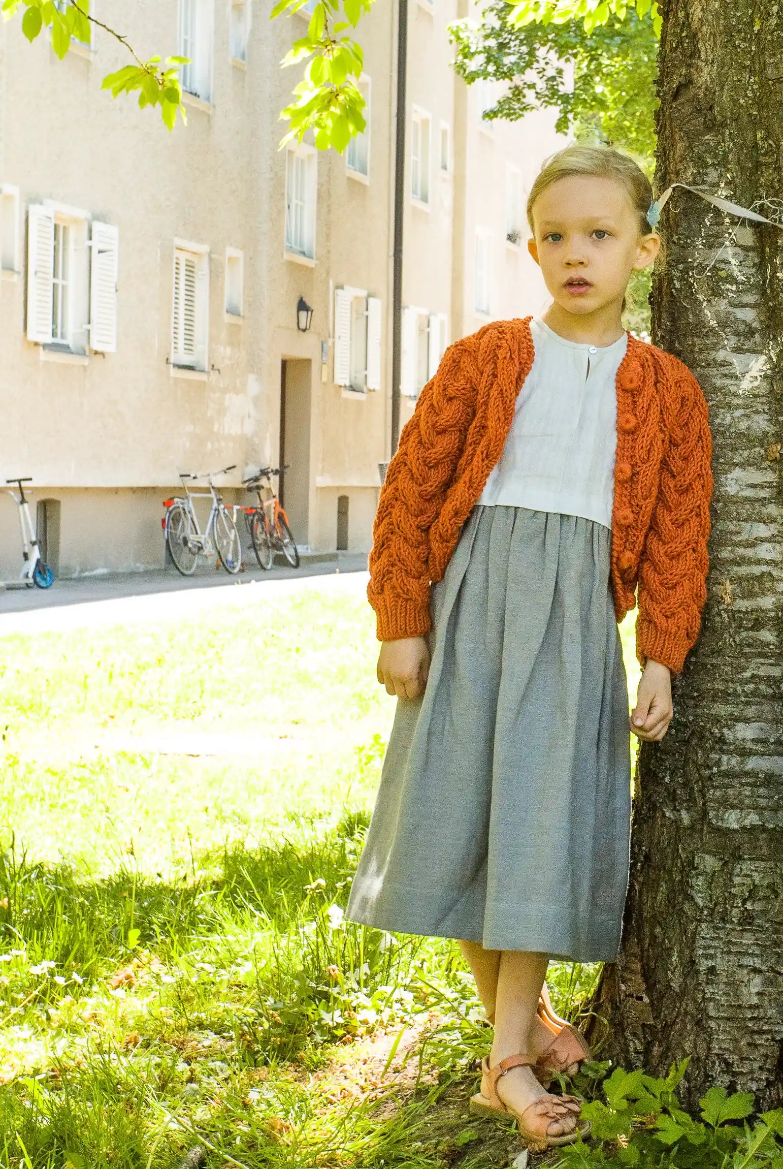 Zirbenfüxlein Kinderdirndl Saphir mit terrakottafarbener Strickjacke, Kind in Frontansicht auf einer Wiese an einen Baum angelehnt in urbaner Hinterhofszene, klar gestaltet von Zirbenfüxlein.