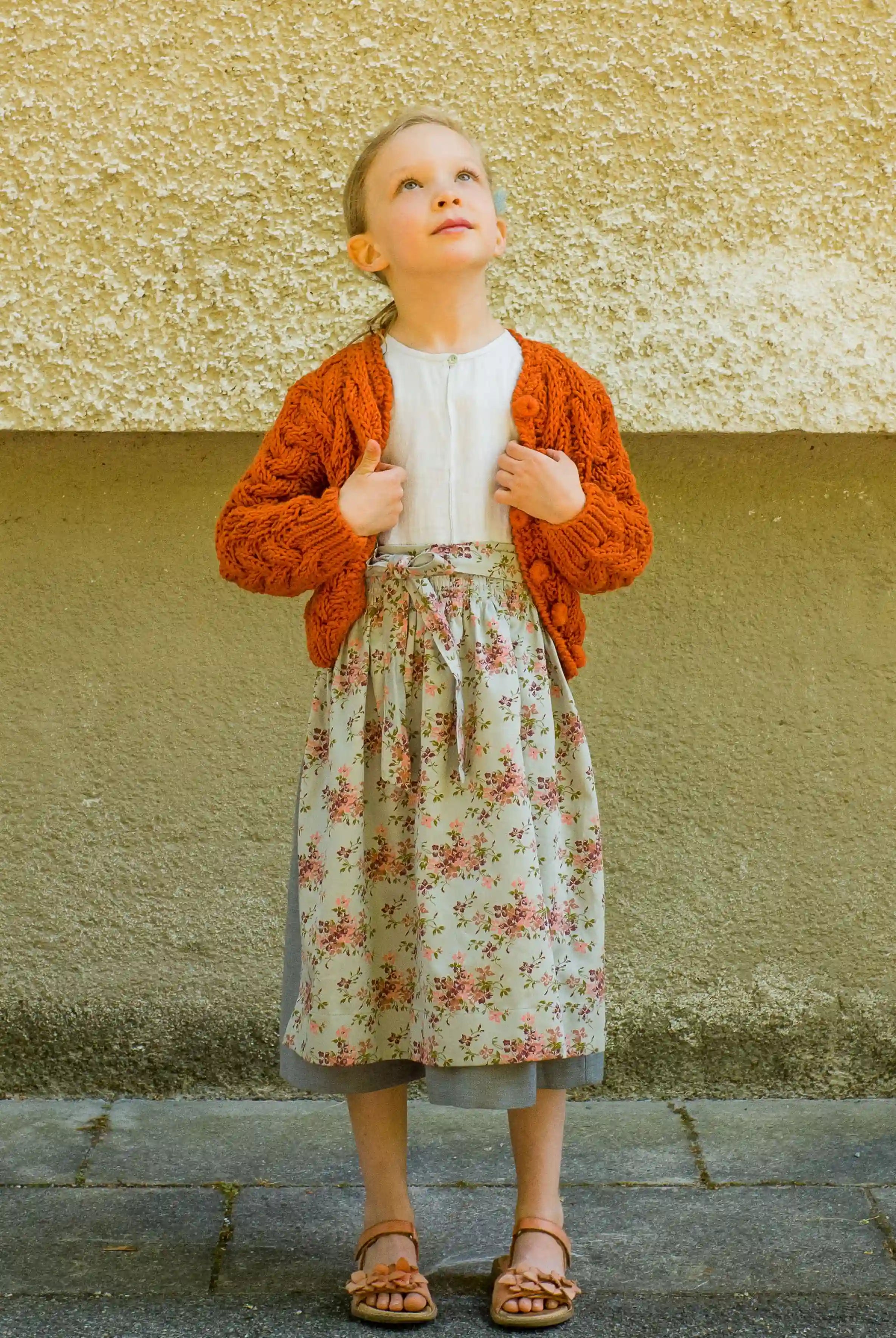 Zirbenfüxlein Kinderdirndl Saphir mit terrakottafarbener Strickjacke und hellblauer Schürze mit Blumenmuster, Frontansicht vor neutraler urbaner Fassade, klar interpretiert von Zirbenfüxlein.