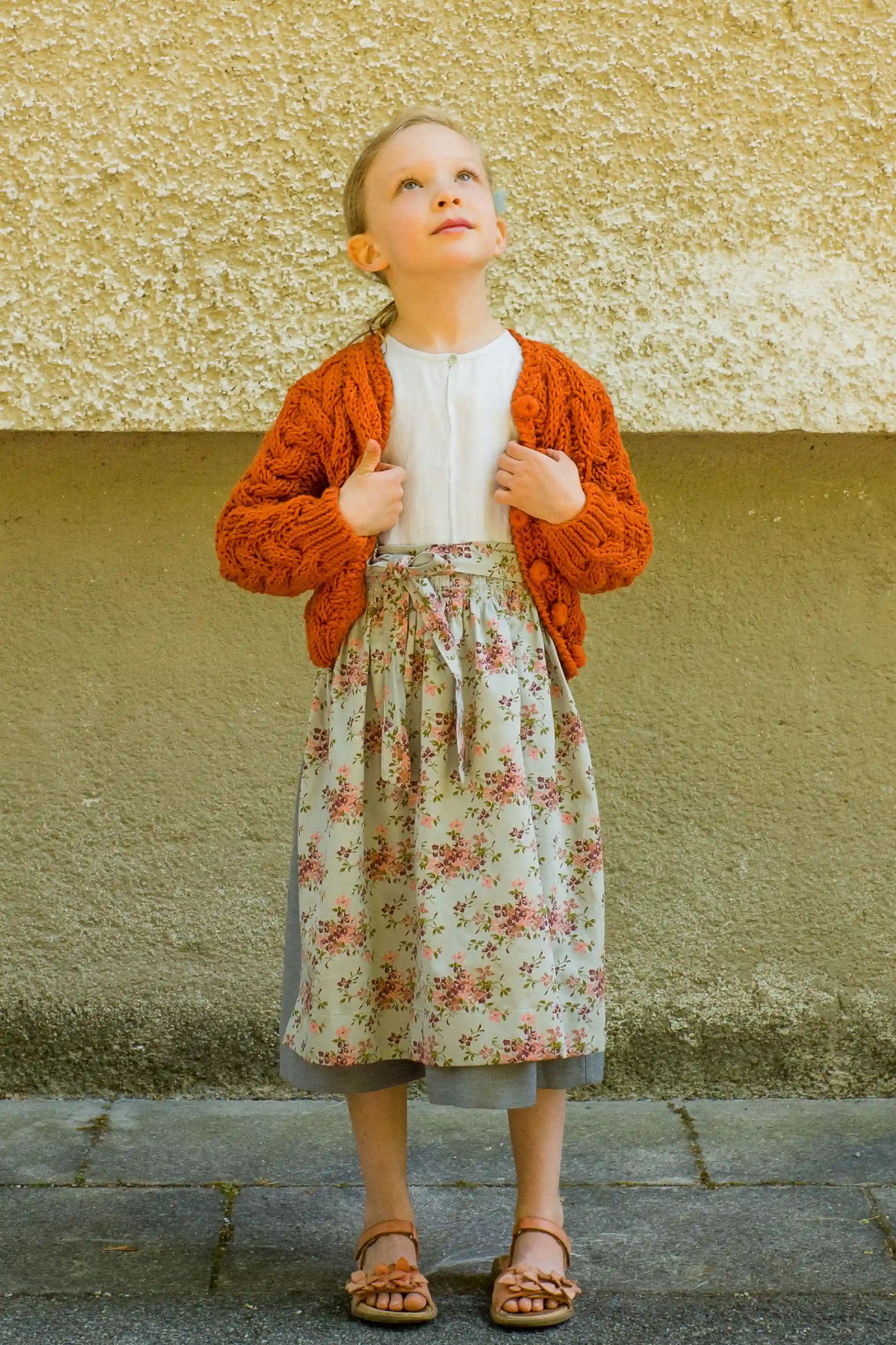 Zirbenfüxlein Kinderdirndl Saphir mit terrakottafarbener Strickjacke und hellblauer Schürze mit Blumenmuster, Frontansicht vor neutraler urbaner Fassade, klar interpretiert von Zirbenfüxlein.