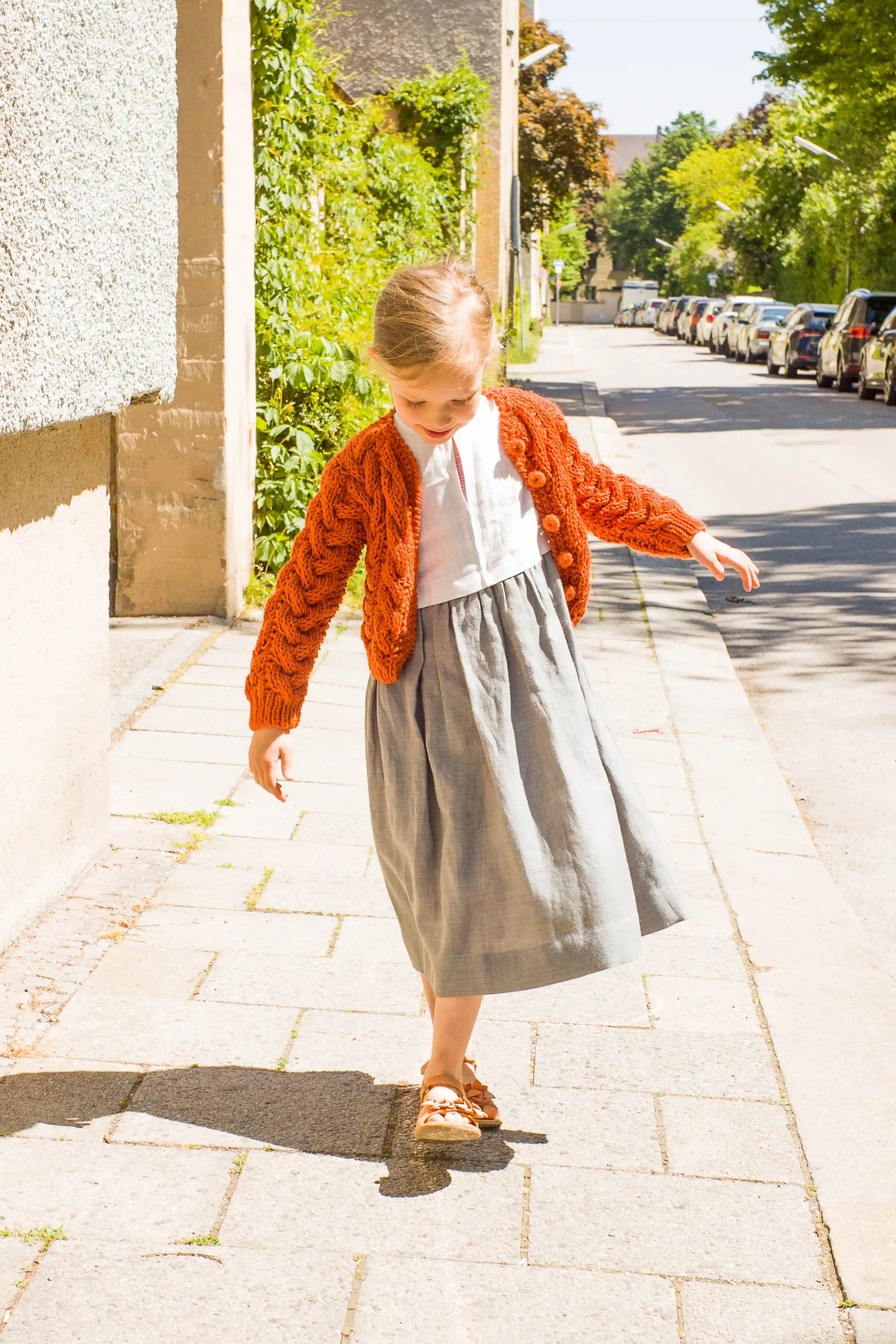 Zirbenfüxlein Kinderdirndl Saphir mit terrakottafarbener Zopf-Strickjacke, Frontansicht eines balancierenden Kindes auf Kreidelinie mit Blick nach unten in sonniger urbaner Straßenszene, authentisch umgesetzt von Zirbenfüxlein.