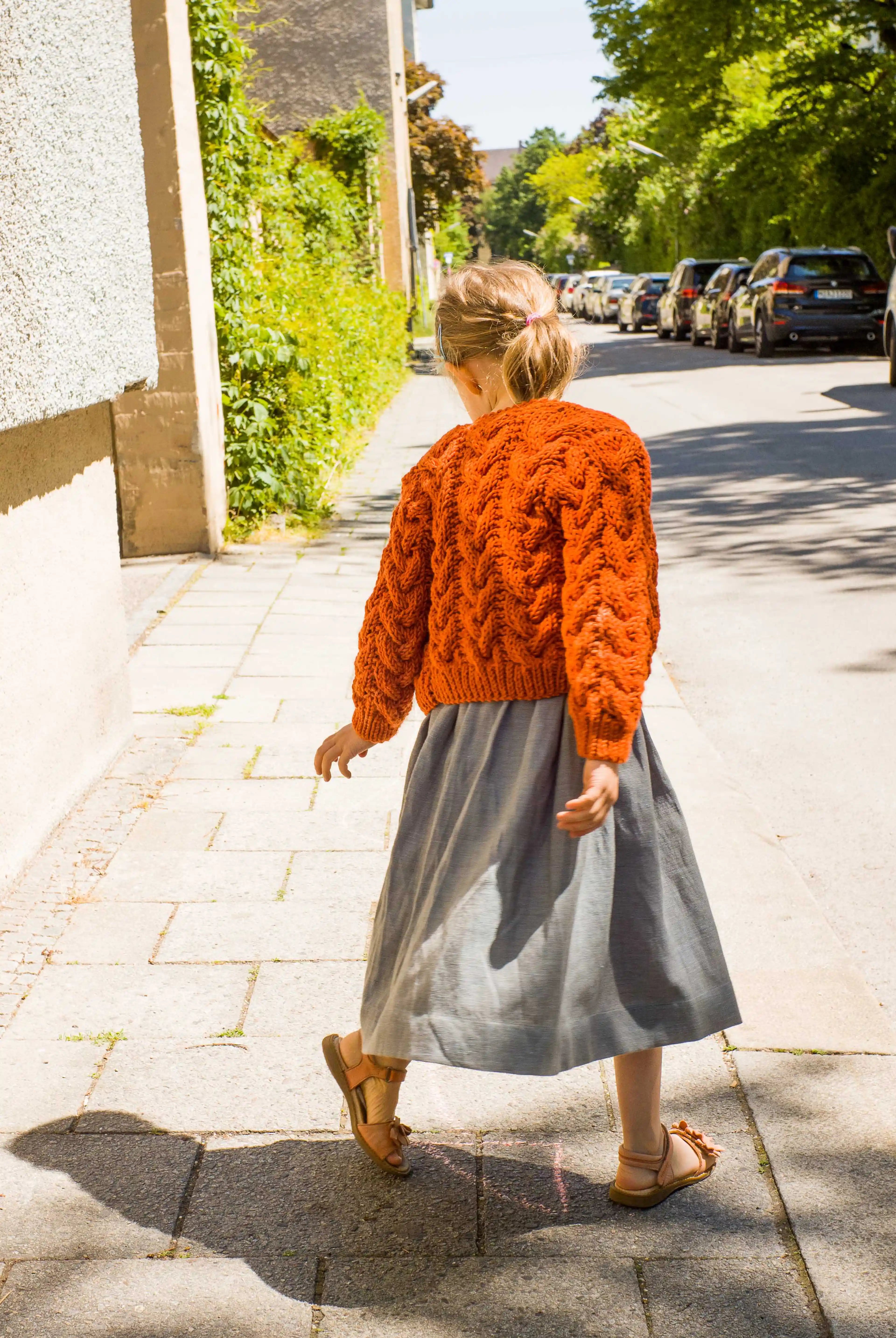 Zirbenfüxlein Kinderdirndl Saphir mit terrakottafarbener Zopf-Strickjacke, Rückansicht eines Kindes in Drehbewegung mit Blick nach unten auf dem Gehsteig in sonniger urbaner Straßenszene, modern interpretiert von Zirbenfüxlein.