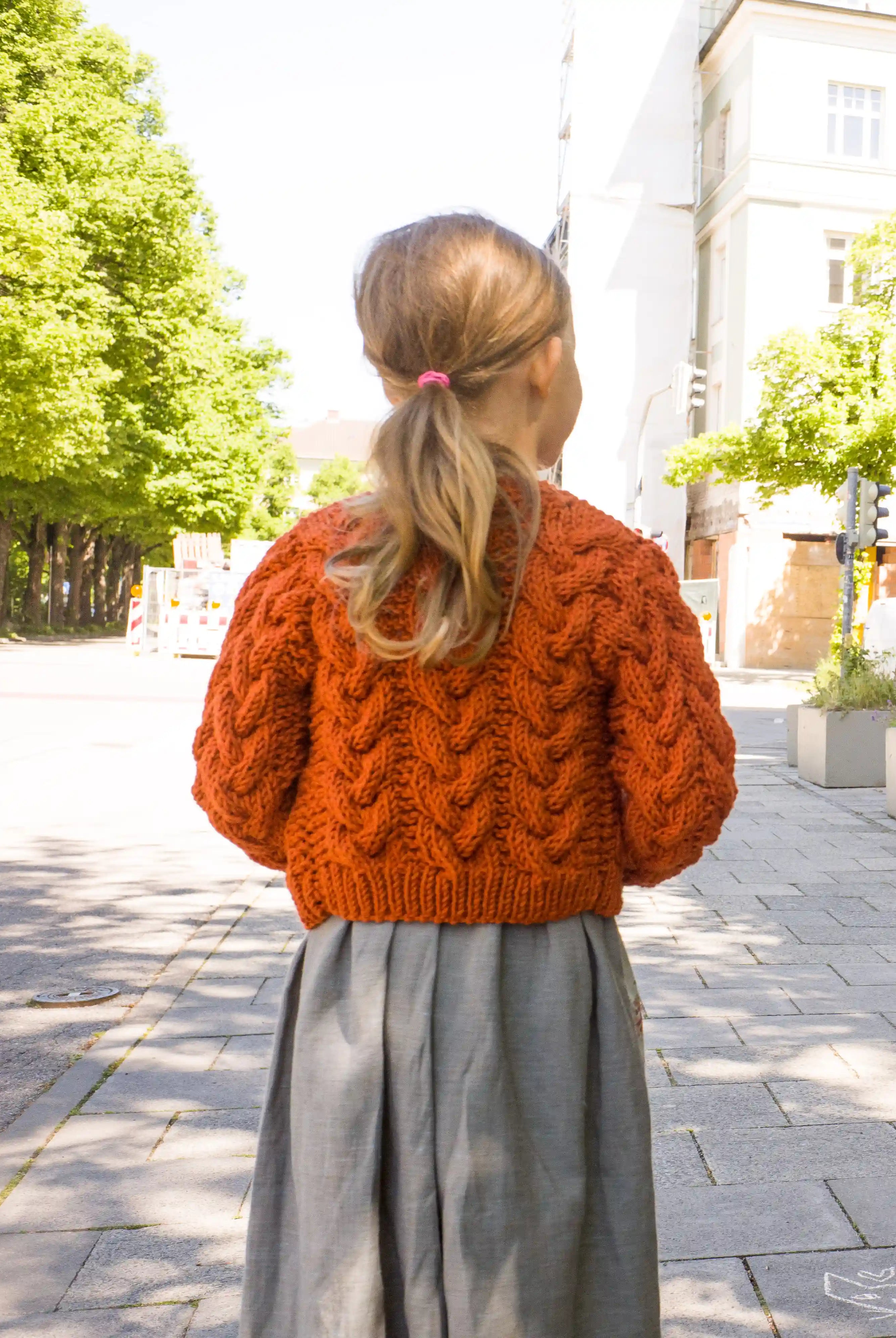 Zirbenfüxlein Kinderdirndl Saphir mit terrakottafarbener Zopf-Strickjacke, Nahaufnahme der Strickjacke in Rückansicht vor urbaner Straßenszene, hochwertig umgesetzt von Zirbenfüxlein.