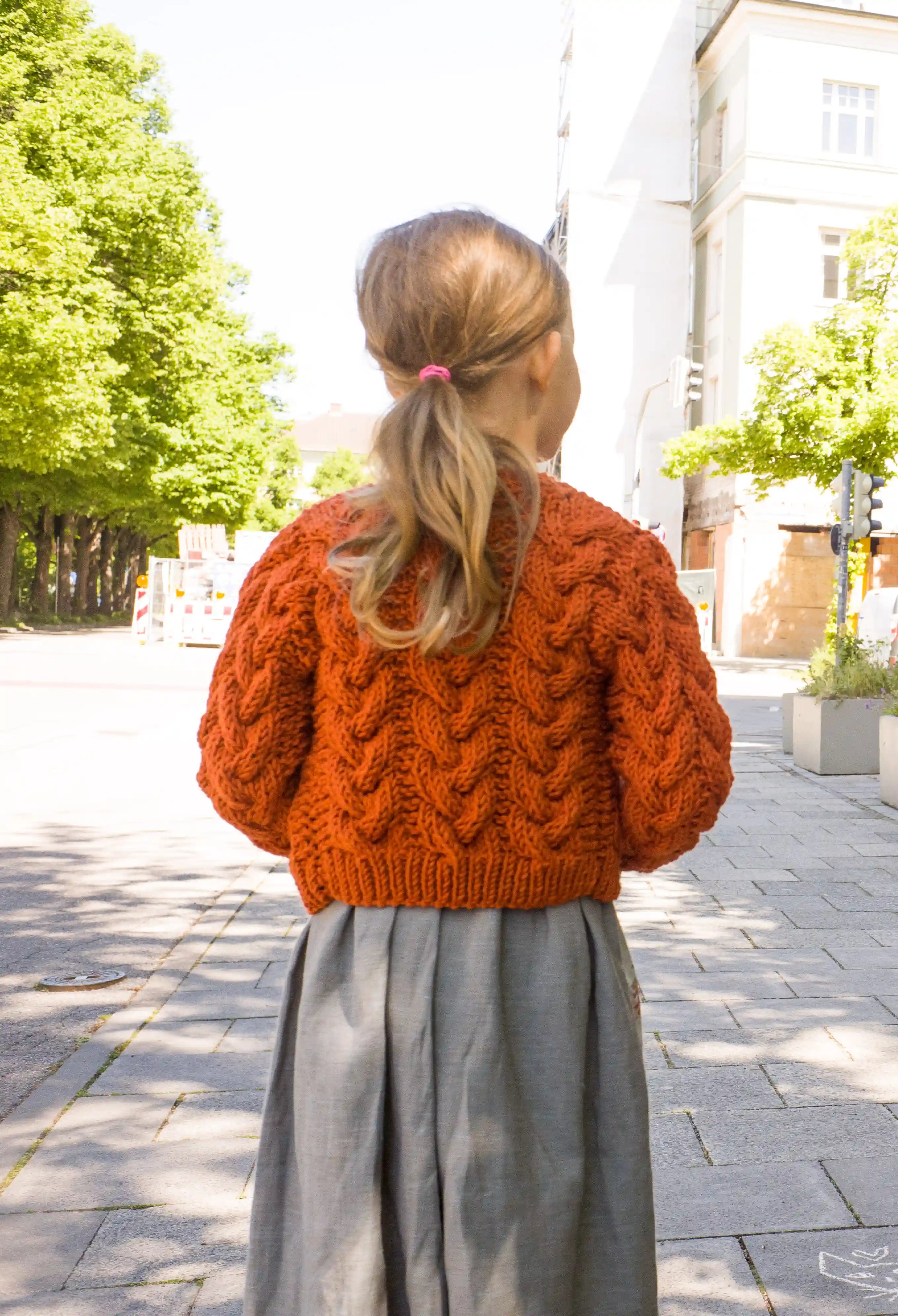 Zirbenfüxlein Kinderdirndl Saphir mit terrakottafarbener Zopf-Strickjacke, Nahaufnahme der Strickjacke in Rückansicht vor urbaner Straßenszene, hochwertig umgesetzt von Zirbenfüxlein.