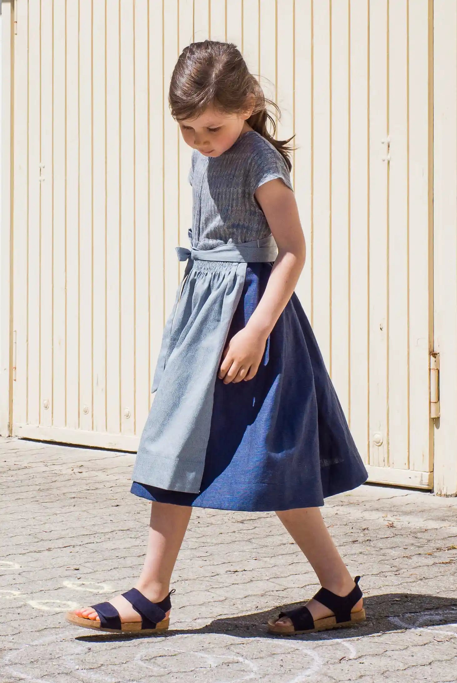 Leinen-Kinderdirndl in Tintenblau mit hellblauer Baumwollschürze – Drehbewegung nach vorne vor Garageneinfahrt, schwungvolle Szene mit Blickrichtung nach unten, lebendig interpretiert von Zirbenfüxlein