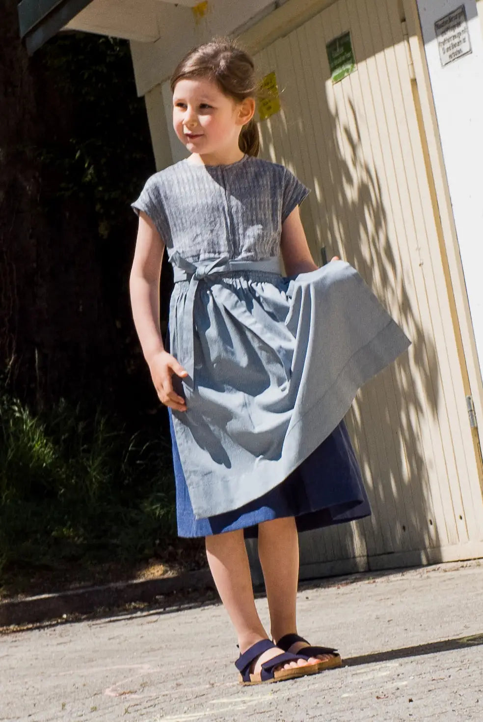 Leinen-Kinderdirndl in Tintenblau mit hellblauer Baumwollschürze – stehend vor Garagentor, das Kind hält die Schürze in der Hand, inszeniert von Zirbenfüxlein