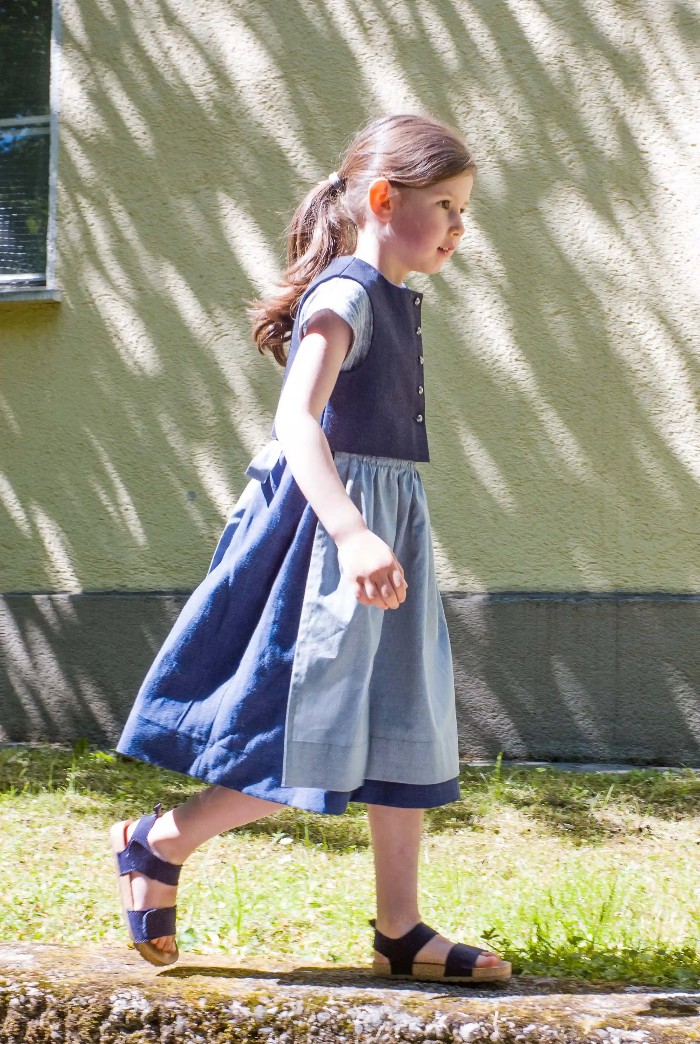 Kinderdirndl in Tintenblau mit hellblauer Schürze – Kind läuft von links nach rechts auf einem Mäuerchen entlang, vor urbaner Häuserfassade mit Licht- und Schattenspiel, lebendig inszeniert von Zirbenfüxlein
