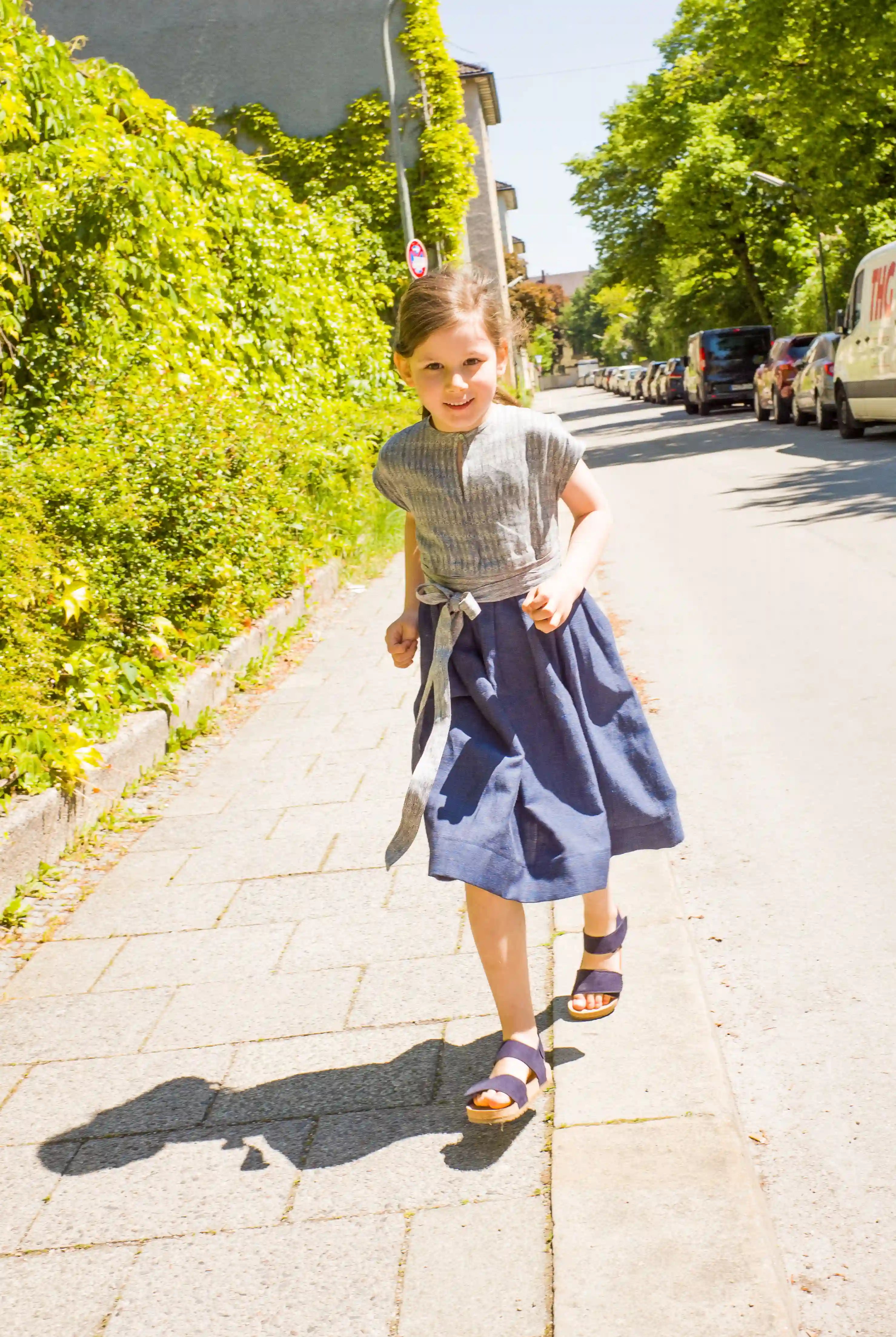 Kinderdirndl in Tintenblau mit Schürzenband – Kind läuft auf Gehweg entlang einer Straße im urbanen Setting, lebendig inszeniert von Zirbenfüxlein
