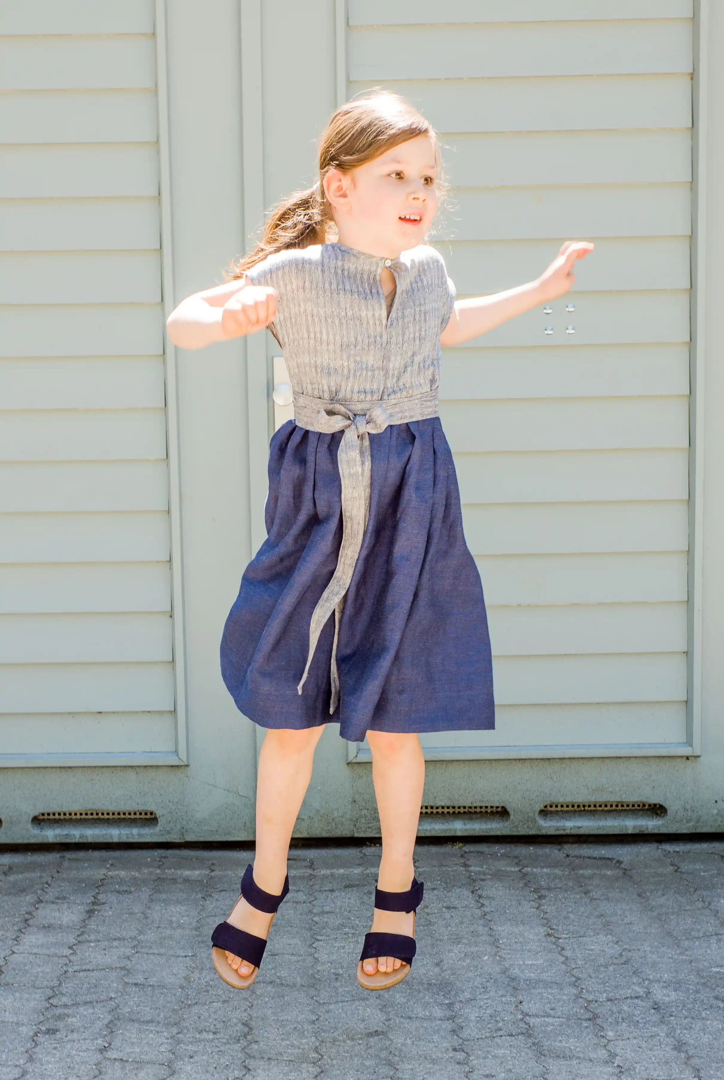 Kinderdirndl in Tintenblau mit Schürzenband – Kind springt vor einem Garagentor nach oben, fröhlich inszeniert von Zirbenfüxlein