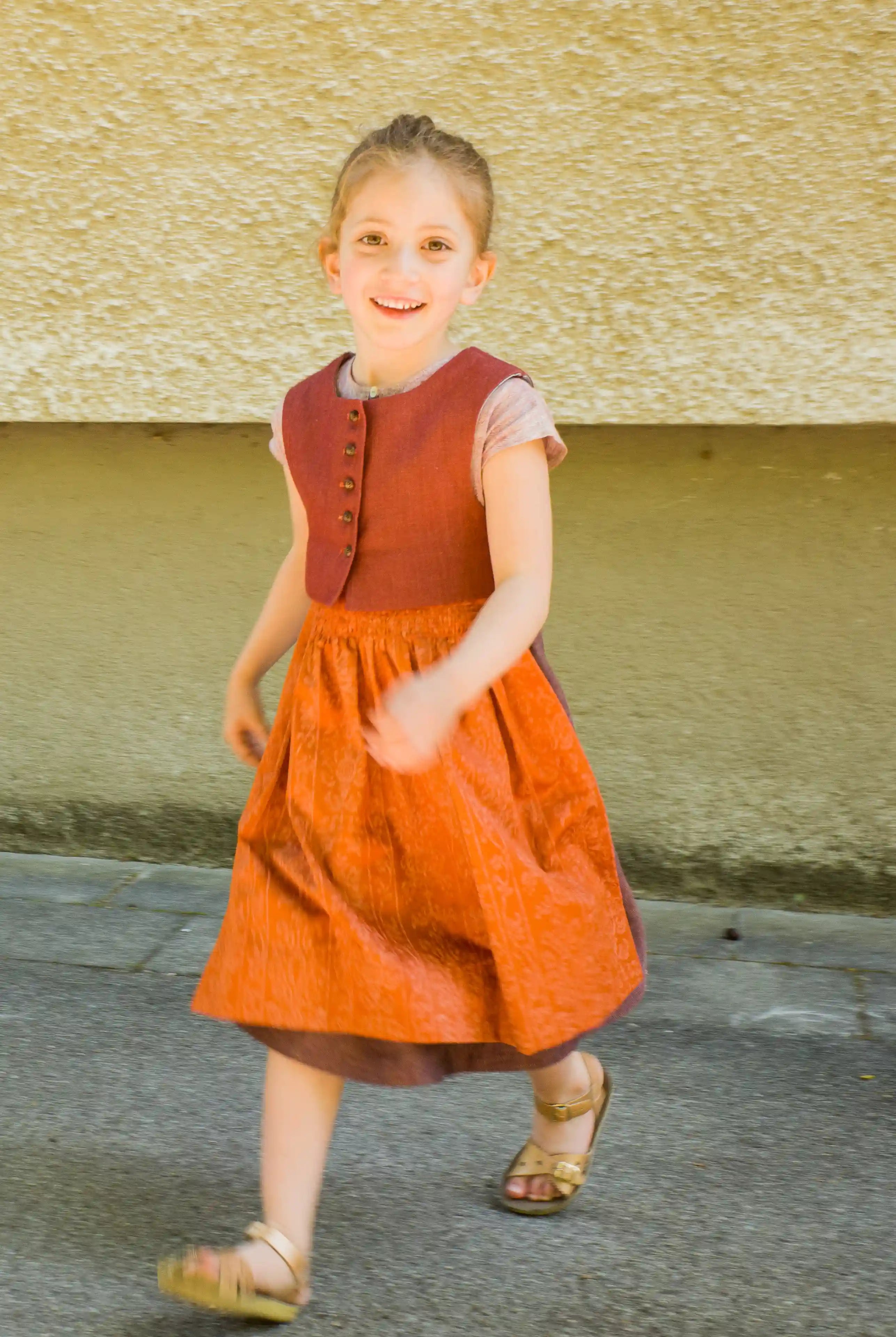Kinderdirndl in pinker Beere mit terrakottafarbener Schürze und Weste – nach vorne aus dem Bild laufend vor urbaner Fassade, lebendig und modern inszeniert von Zirbenfüxlein