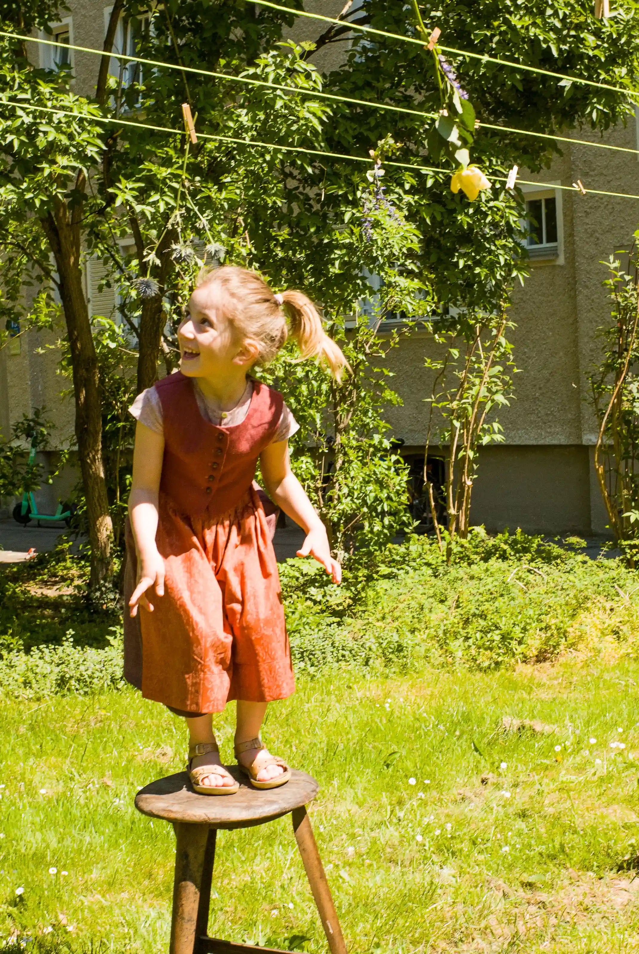 Kinderdirndl in Pink mit terrakottafarbener Schürze und Weste – in einer Wiese in einem Hinterhof auf einem alten Hocker stehend, Blick nach oben zu einer Wäscheleine, inszeniert von Zirbenfüxlein