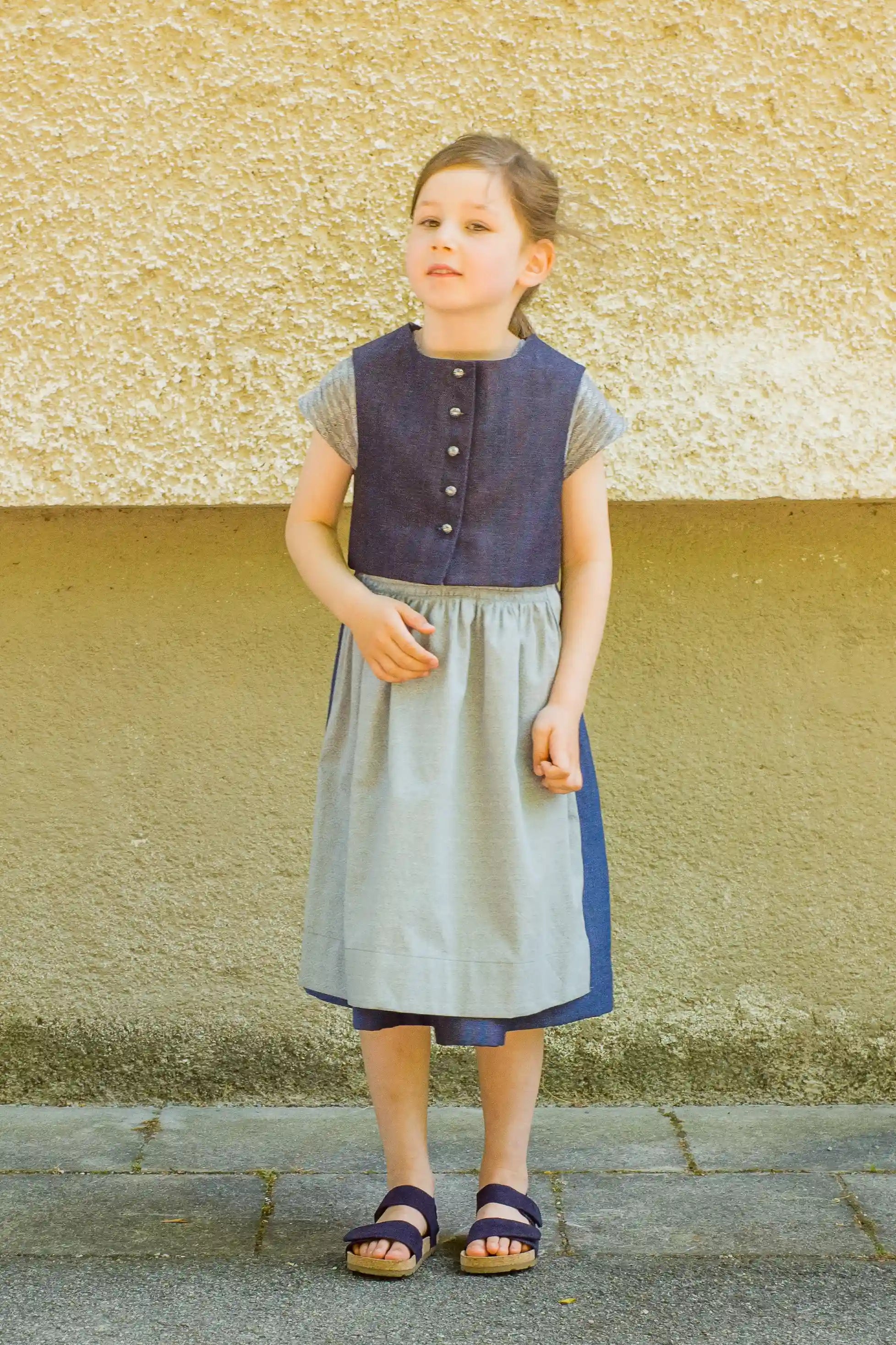 Kinderdirndl mit Weste in Tintenblau und hellblauer Schürze – Frontansicht vor neutraler Fassadenwand, klassisch und reduziert inszeniert von Zirbenfüxlein