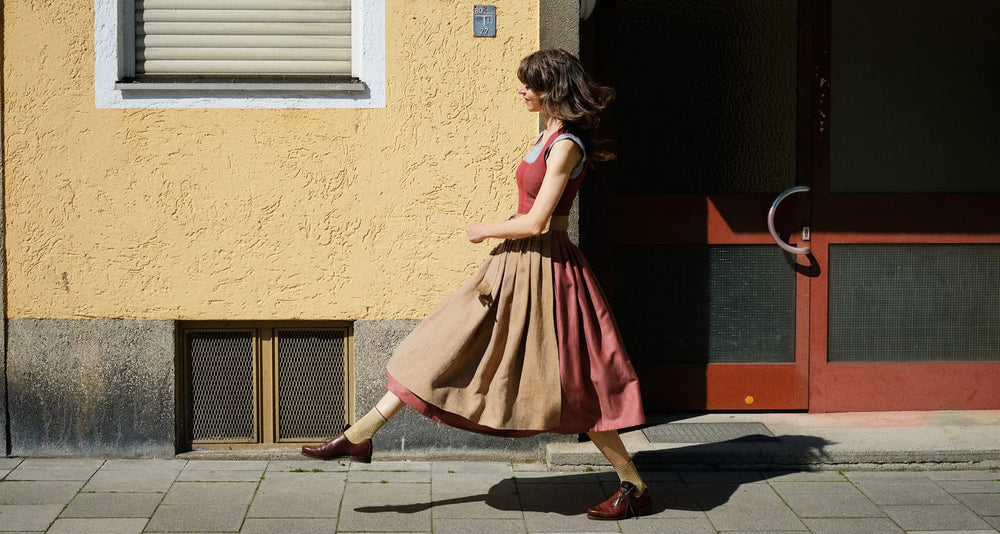 Model trägt Dirndl Ava in pinker Beere mit ocker-gestreifter Schürze – läuft schwungvoll von rechts nach links über den Gehweg vor urbaner Häuserfassade, moderne Tracht lebendig in Szene gesetzt von Zirbenfux
