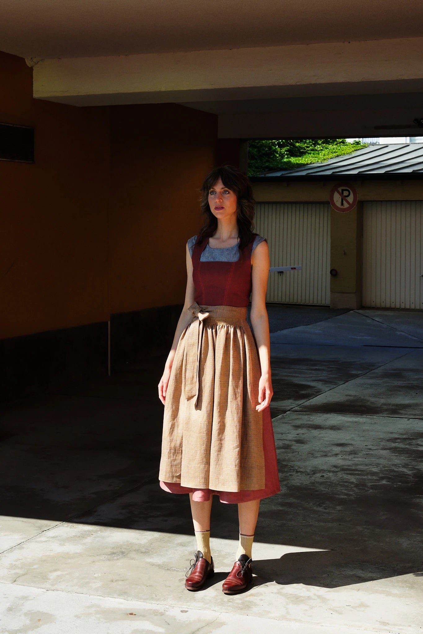 Model trägt Dirndl Ava in pinker Beere mit ocker-gestreifter Schürze – steht regungslos in einer Garageneinfahrt, halb im Schatten, halb in der Sonne, urban-minimalistisch inszeniert von Zirbenfux