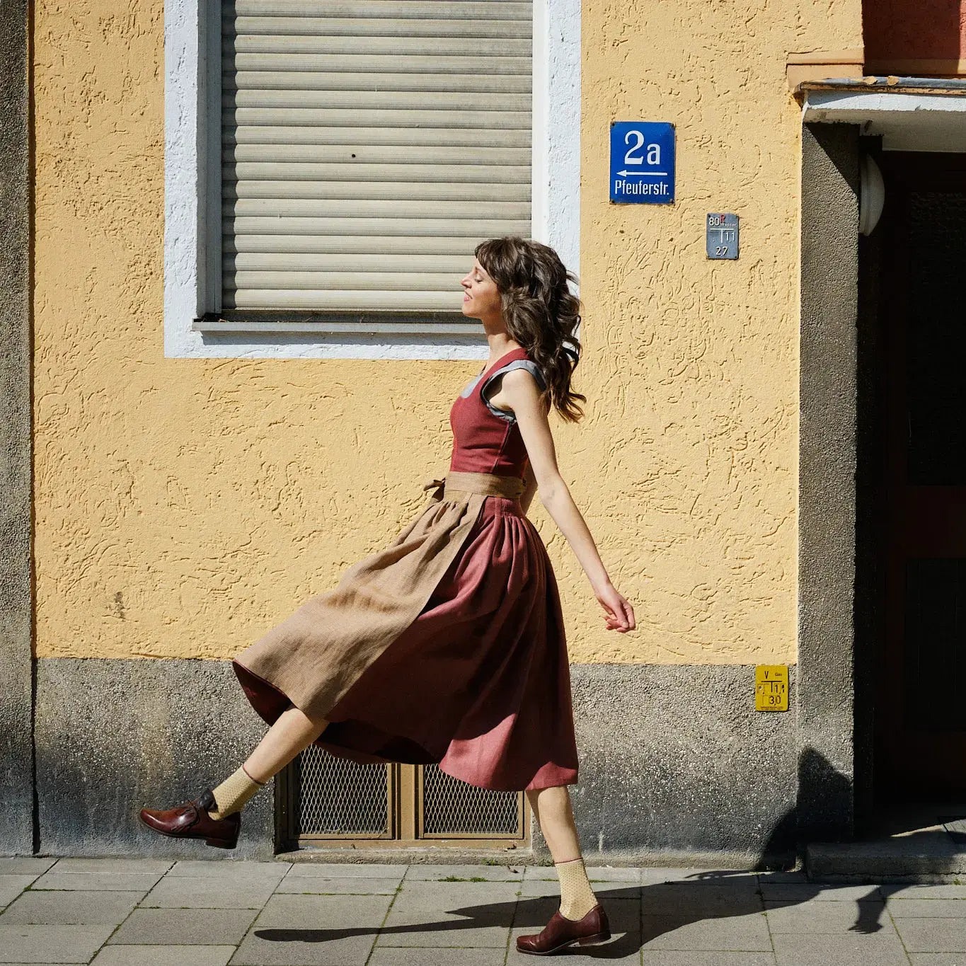 Dirndl Ava in Pinke Beere aus Leinen mit ocker gestreifter Schürze, Model läuft schwungvoll auf dem Gehweg vor urbaner Häuserfassade von rechts nach links durch das Bild, dynamisch inszeniert von Zirbenfux.