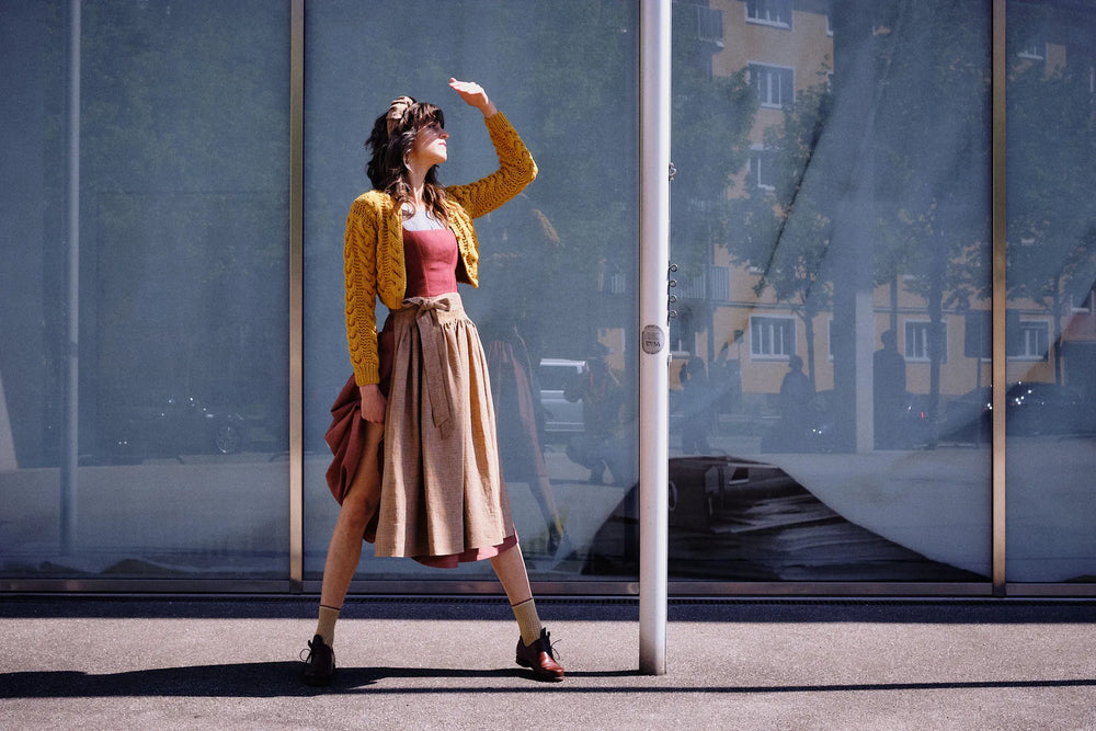 Dirndl Ava in Pinke Beere aus Leinen mit ocker gestreifter Schürze und Strickjacke, Model im Profil vor moderner Glasfassade mit Blick zur Sonne, eine Hand schützend vor dem Gesicht, die andere rafft den Rock am Bein entlang, klar inszeniert von Zirbenfux.