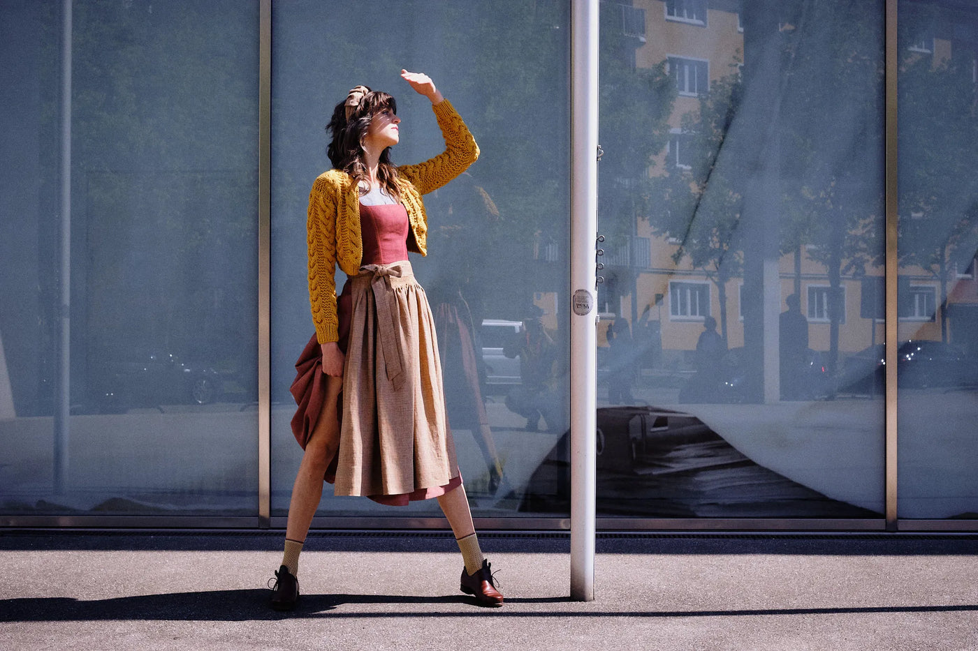 Dirndl Ava in Pinke Beere aus Leinen mit ocker gestreifter Schürze und Strickjacke, Model im Profil vor moderner Glasfassade mit Blick zur Sonne, eine Hand schützend vor dem Gesicht, die andere rafft den Rock am Bein entlang, klar inszeniert von Zirbenfux.