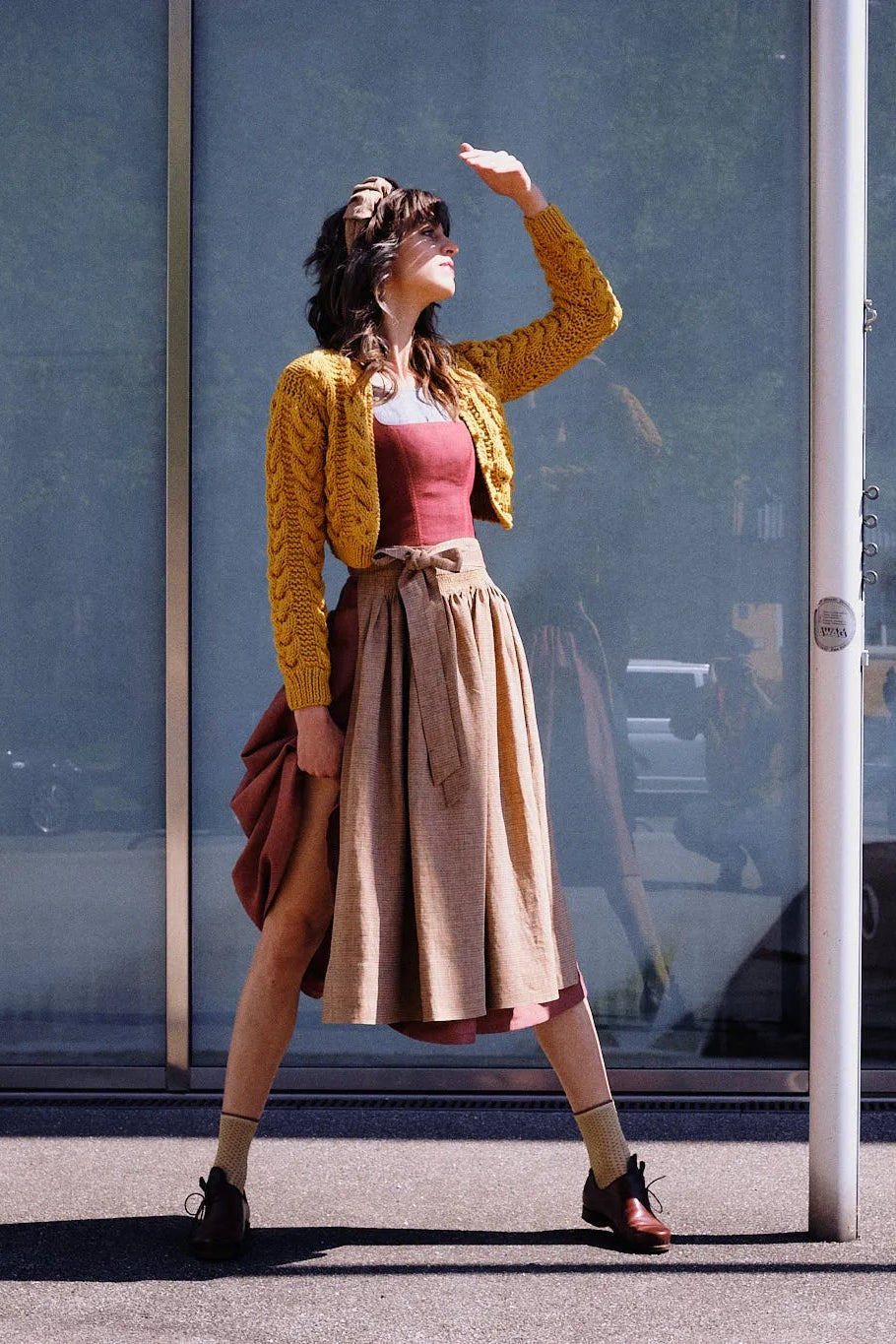 Dirndl Ava in Pinke Beere aus Leinen mit ocker gestreifter Schürze und Strickjacke, Model im Profil vor moderner Glasfassade mit Blick zur Sonne, eine Hand schützend vor dem Gesicht, die andere rafft den Rock am Bein entlang, klar inszeniert von Zirbenfux.