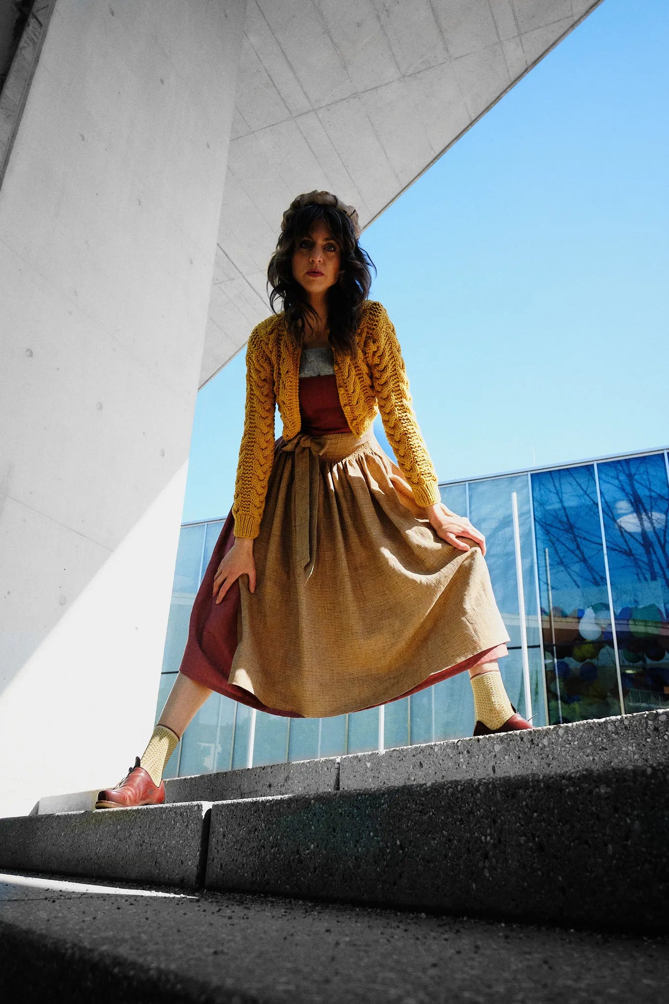 Dirndl Ava in Pinke Beere aus Leinen mit ocker gestreifter Schürze und Strickjacke, Model vor moderner Fassade nach vorne zur Kamera gebeugt von unten fotografiert, direkter Blick in die Kamera, spannungsvoll inszeniert von Zirbenfux.