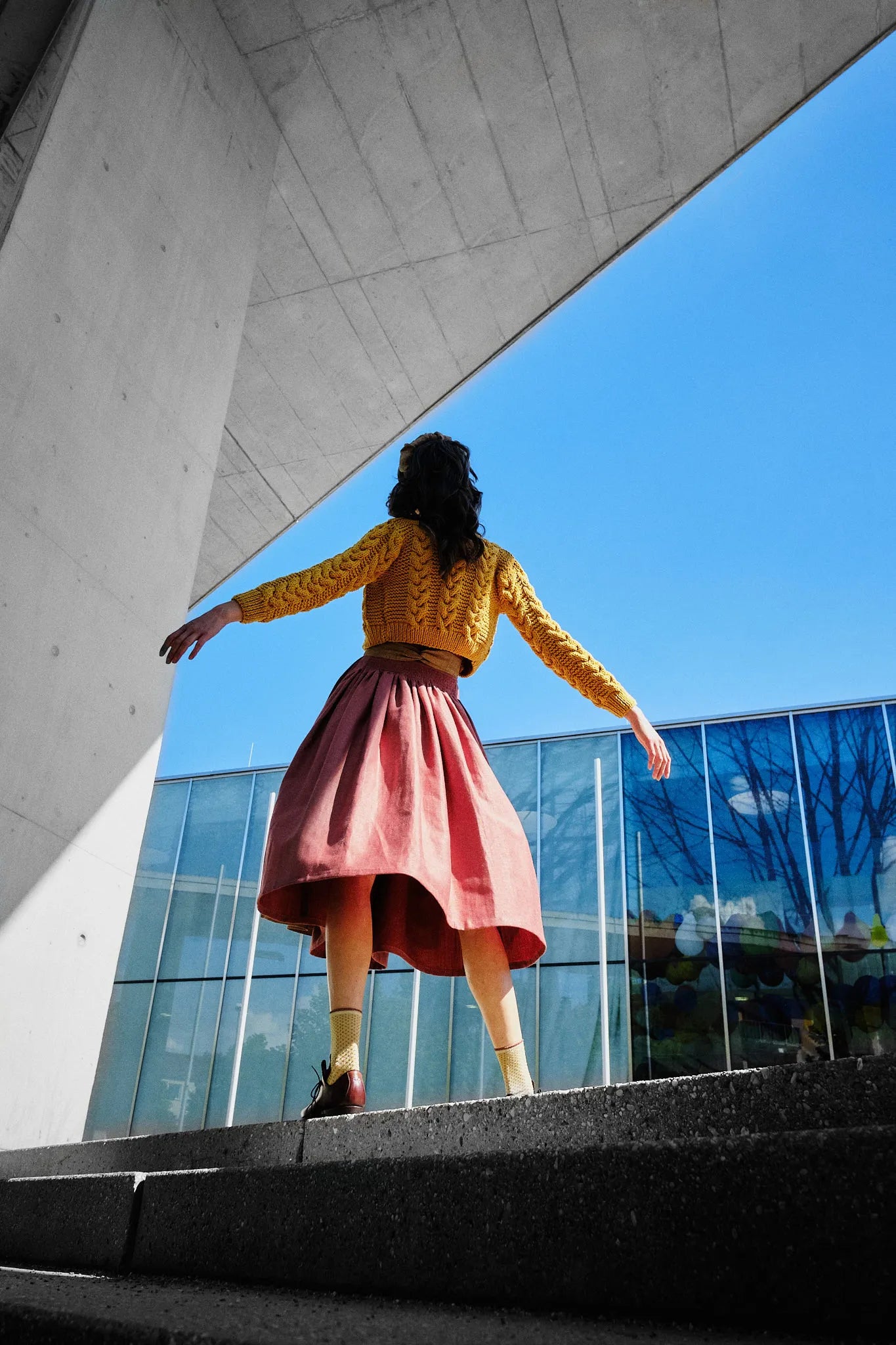 Dirndl Ava in Pinke Beere aus Leinen mit ocker gestreifter Schürze und Strickjacke, Rückansicht mit Perspektive vor moderner Fassade an Treppenabsatz mit seitlich ausgestreckten Armen und weit schwingendem Rock, kraftvoll inszeniert von Zirbenfux.