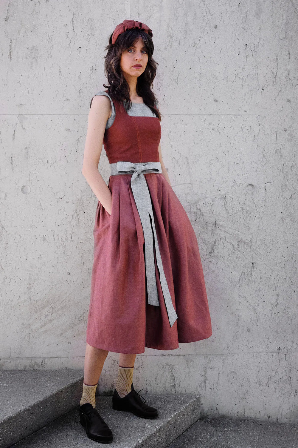 Dirndl Ava in Pinke Beere aus Leinen mit blaugrauem Schürzenband, Model klar und gerade vor einer modernen Waschbetonwand stehend, Hände locker in den Rocktaschen, gelassener Blick in die Kamera, ruhig inszeniert von Zirbenfux.