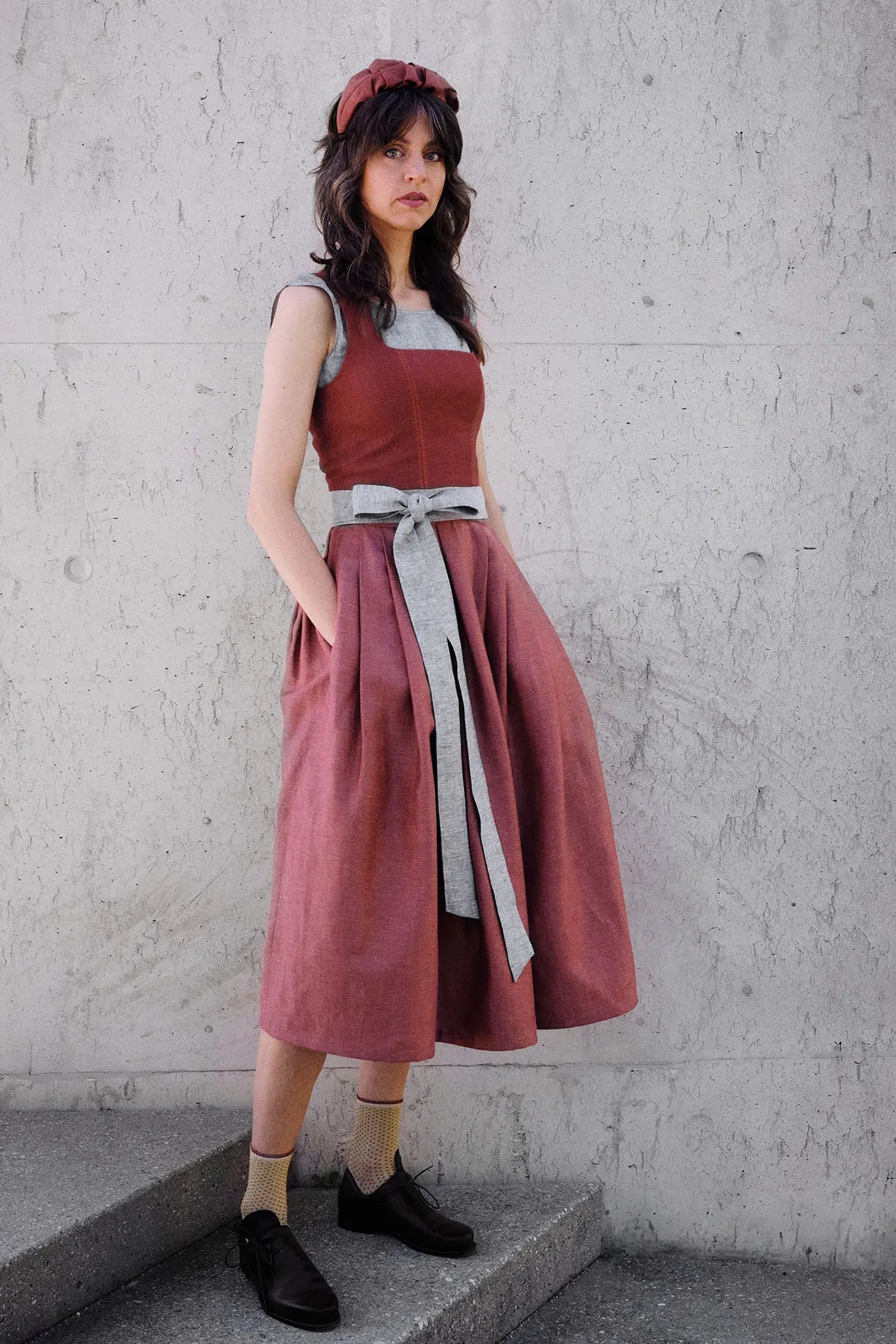 Dirndl Ava in Pinke Beere aus Leinen mit blaugrauem Schürzenband, Model klar und gerade vor einer modernen Waschbetonwand stehend, Hände locker in den Rocktaschen, gelassener Blick in die Kamera, ruhig inszeniert von Zirbenfux.
