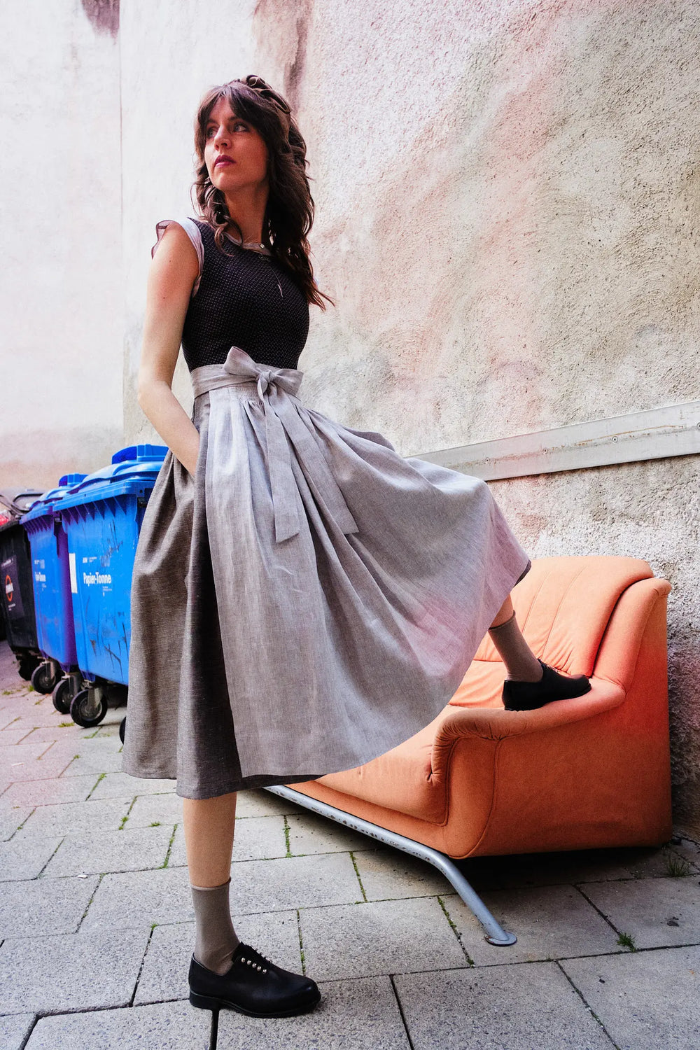 Model trägt Dirndl Marlene in Schwarz-Natur mit Leinenschürze – steht in urbaner Hinterhofszenerie mit einem Bein auf einem ausgeblichenen orangenen Sofa, Hände lässig in den Rocktaschen, Blick zurück über die Schulter, modern interpretiert von Zirbenfux